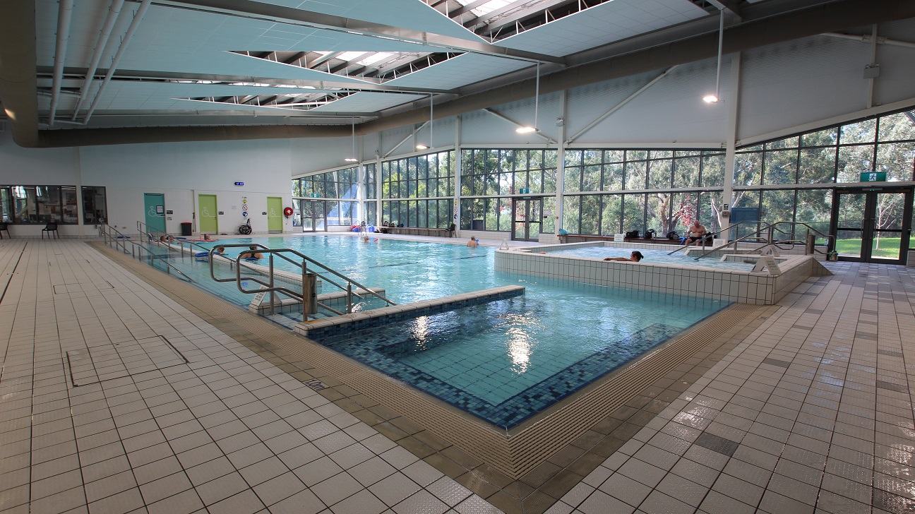 Indoor swimming pool with a tiled deck surrounding it. The pool has metal handrails and a few people are in the water. Large windows on the right side provide a view of trees outside. The ceiling is high with exposed beams and skylights. The floor is covered with light-coloured tiles. Doors and emergency equipment are visible on the far wall. Indoor swimming pool with a tiled deck surrounding it. The pool has metal handrails and a few people are in the water. Large windows on the right side provide a view of trees outside. The ceiling is high with exposed beams and skylights. The floor is covered with light-coloured tiles. Doors and emergency equipment are visible on the far wall.