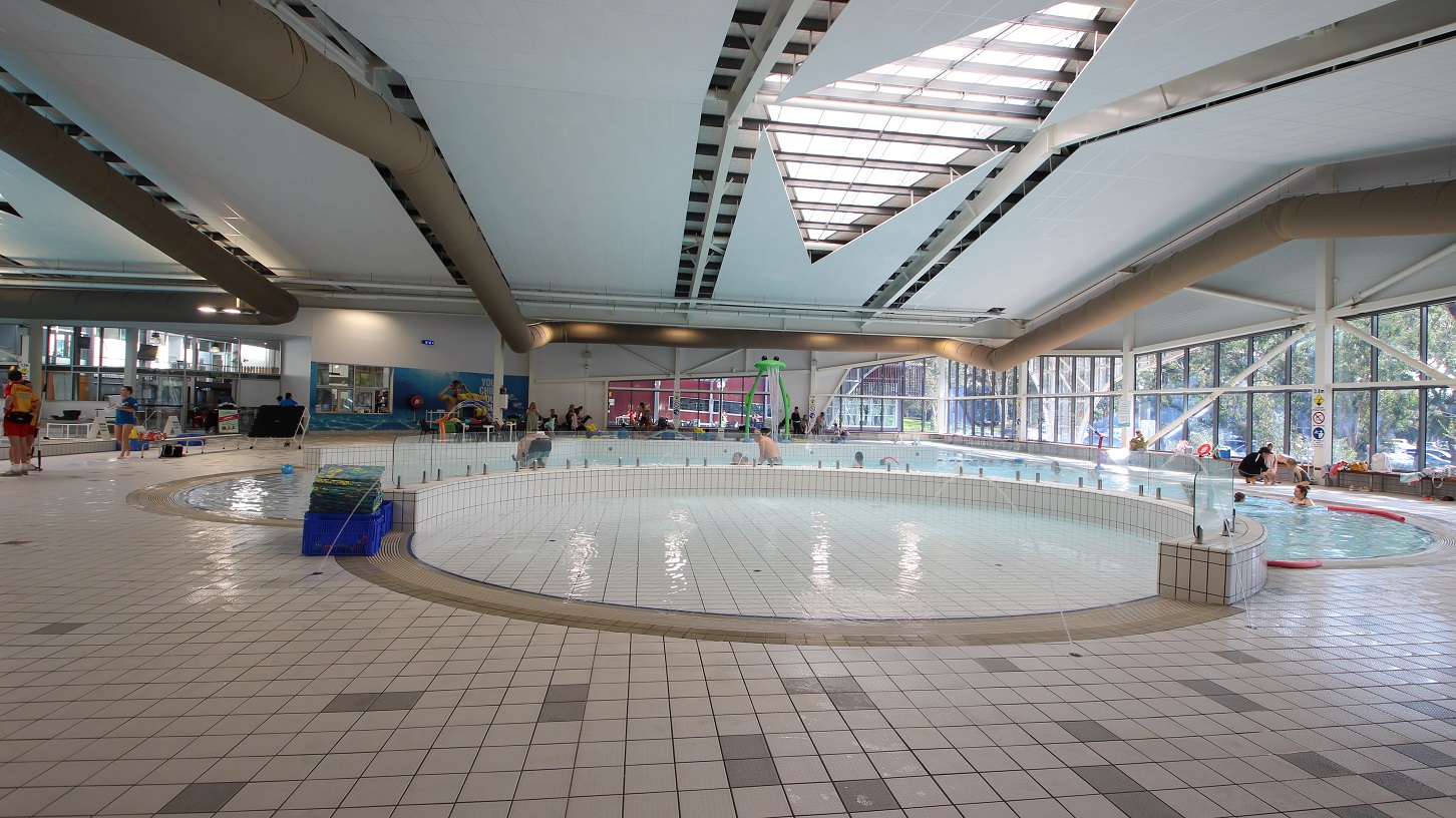 A spacious indoor swimming pool area with a large, circular shallow pool in the centre. The floor is tiled with a mix of beige and grey tiles. On the left, there are people standing near the pool's edge. The right side features large windows allowing natural light to enter, with trees visible outside. The ceiling is high with geometric designs and skylights. In the background, there are more pools and people, along with some poolside equipment. A spacious indoor swimming pool area with a large, circular shallow pool in the centre. The floor is tiled with a mix of beige and grey tiles. On the left, there are people standing near the pool's edge. The right side features large windows allowing natural light to enter, with trees visible outside. The ceiling is high with geometric designs and skylights. In the background, there are more pools and people, along with some poolside equipment.