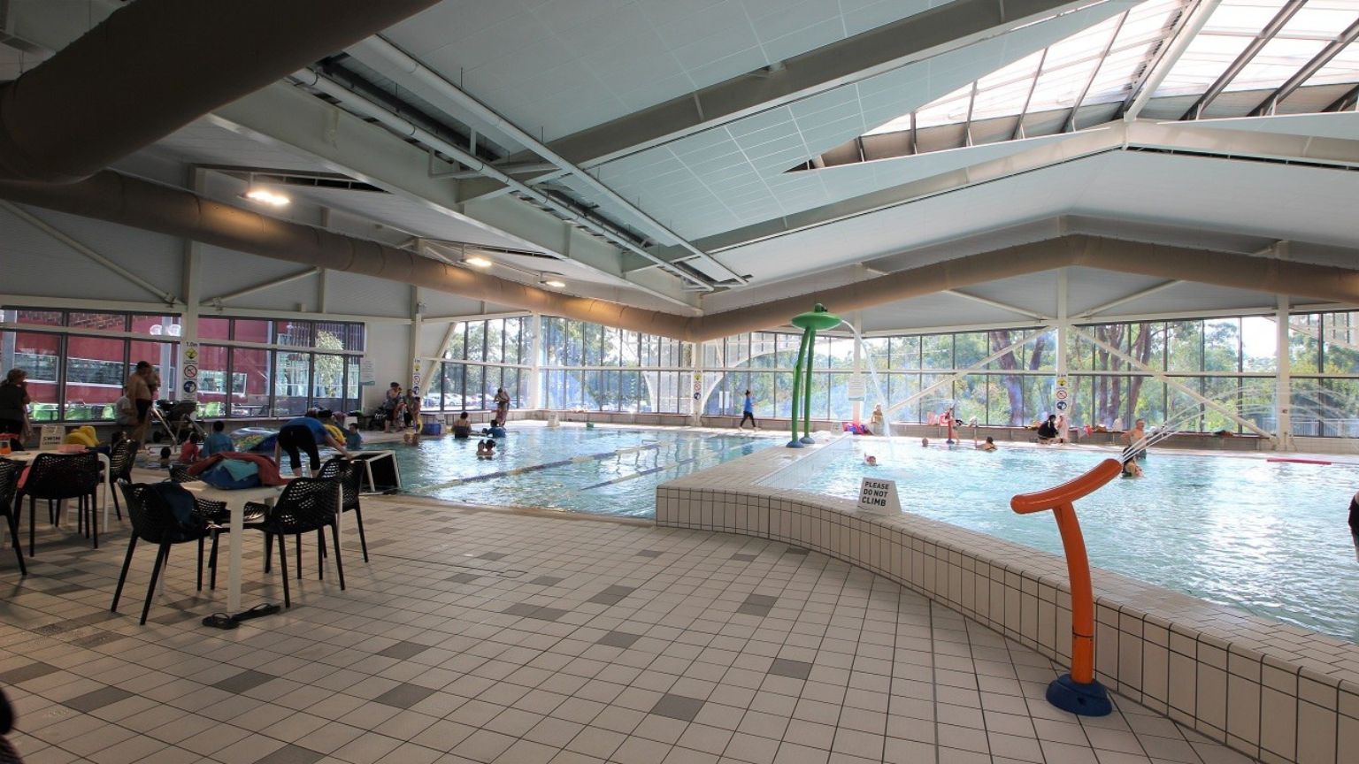 Indoor swimming pool area with a tiled floor. On the left, there are chairs and tables with people sitting and standing nearby. The pool is in the centre, with several people swimming and playing. A green water feature stands in the pool. Large windows in the background allow natural light to enter, and trees are visible outside. The ceiling is high with exposed beams and lighting fixtures. Indoor swimming pool area with a tiled floor. On the left, there are chairs and tables with people sitting and standing nearby. The pool is in the centre, with several people swimming and playing. A green water feature stands in the pool. Large windows in the background allow natural light to enter, and trees are visible outside. The ceiling is high with exposed beams and lighting fixtures.
