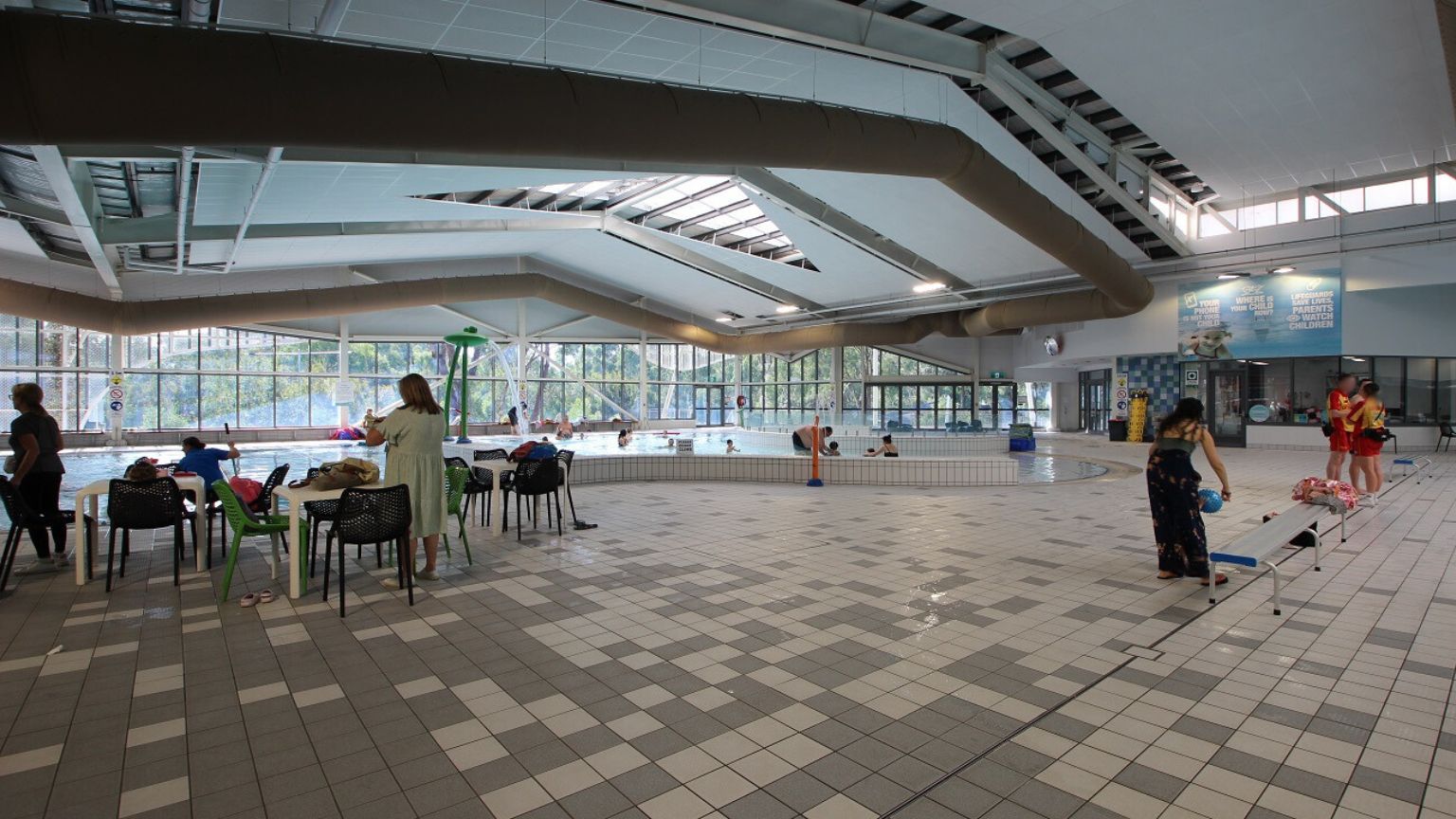 Indoor swimming pool area with a tiled floor in shades of green and beige. On the left, several people are seated at tables with chairs, some watching the pool. In the centre, a pool with swimmers and a lifeguard is visible. On the right, a person is bending over near a small poolside structure, and a group of people are gathered near the wall. The ceiling has a geometric design with skylights, and large windows in the background allow natural light in. Indoor swimming pool area with a tiled floor in shades of green and beige. On the left, several people are seated at tables with chairs, some watching the pool. In the centre, a pool with swimmers and a lifeguard is visible. On the right, a person is bending over near a small poolside structure, and a group of people are gathered near the wall. The ceiling has a geometric design with skylights, and large windows in the background allow natural light in.