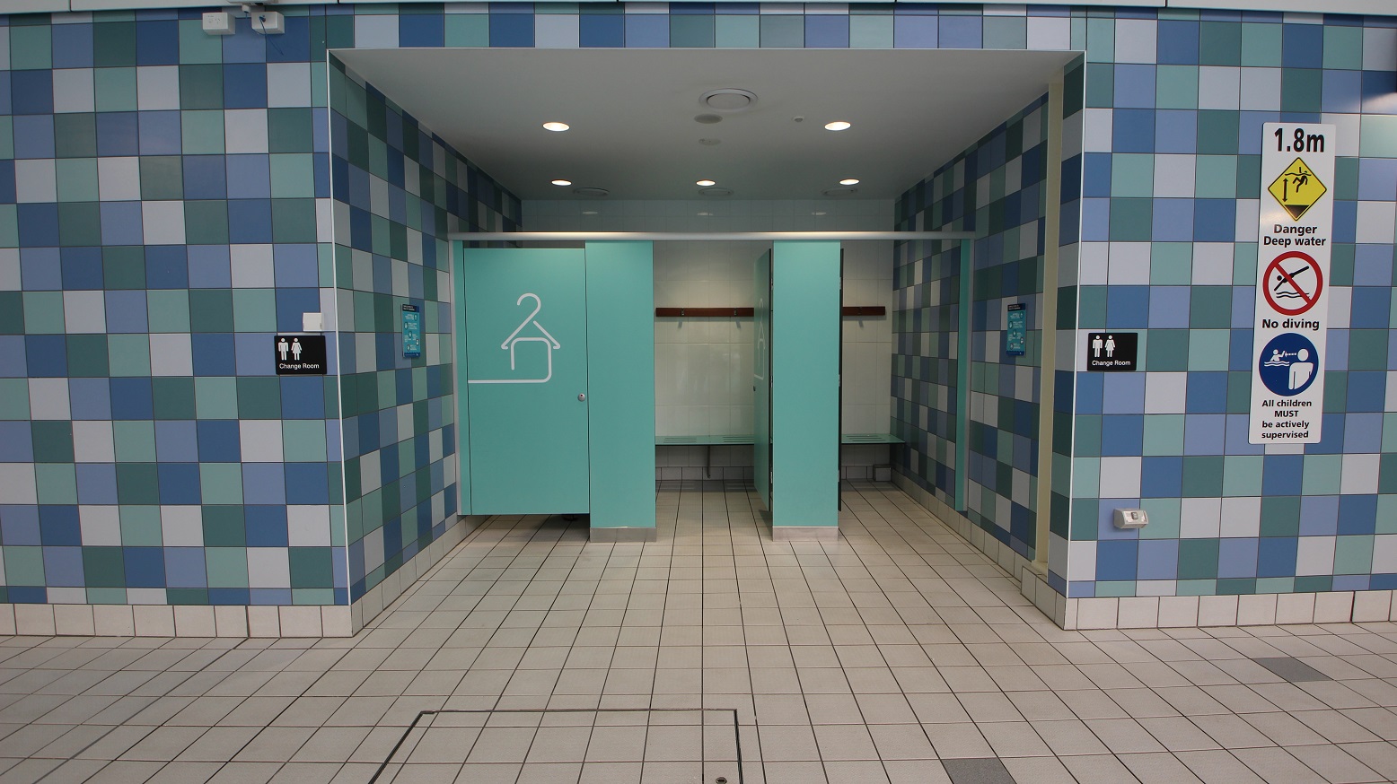 A changing room entrance with three turquoise doors, one slightly open, revealing benches and hooks inside. The walls are covered in a blue and white checkered tile pattern. On the right, there are signs indicating a depth warning, no diving, and supervision requirements. The floor is tiled in a light beige colour, and the ceiling has recessed lighting. A changing room entrance with three turquoise doors, one slightly open, revealing benches and hooks inside. The walls are covered in a blue and white checkered tile pattern. On the right, there are signs indicating a depth warning, no diving, and supervision requirements. The floor is tiled in a light beige colour, and the ceiling has recessed lighting.