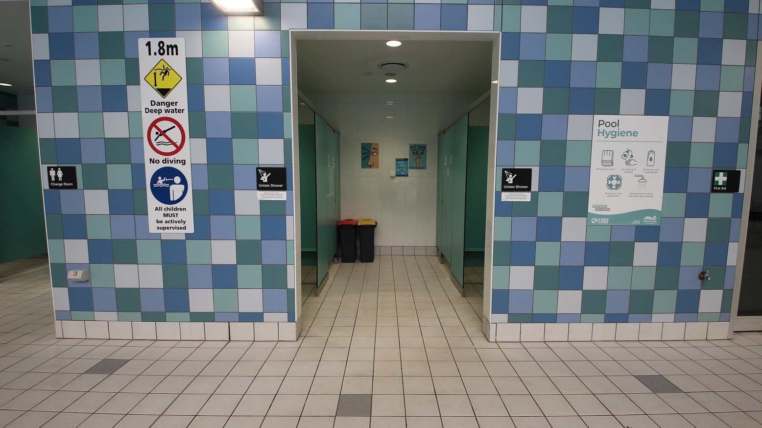 A tiled wall with a blue and white checkerboard pattern. In the centre, there's an entrance to a shower area with two bins inside. Above the entrance, signs indicate "1.8m Danger Deep Water," "No Diving," and "All children MUST be actively supervised." On the left, a sign for the "Change Room" and on the right, a "Pool Hygiene" poster and a "First Aid" sign. The floor is tiled in a light colour, matching the wall's pattern. A tiled wall with a blue and white checkerboard pattern. In the centre, there's an entrance to a shower area with two bins inside. Above the entrance, signs indicate "1.8m Danger Deep Water," "No Diving," and "All children MUST be actively supervised." On the left, a sign for the "Change Room" and on the right, a "Pool Hygiene" poster and a "First Aid" sign. The floor is tiled in a light colour, matching the wall's pattern.