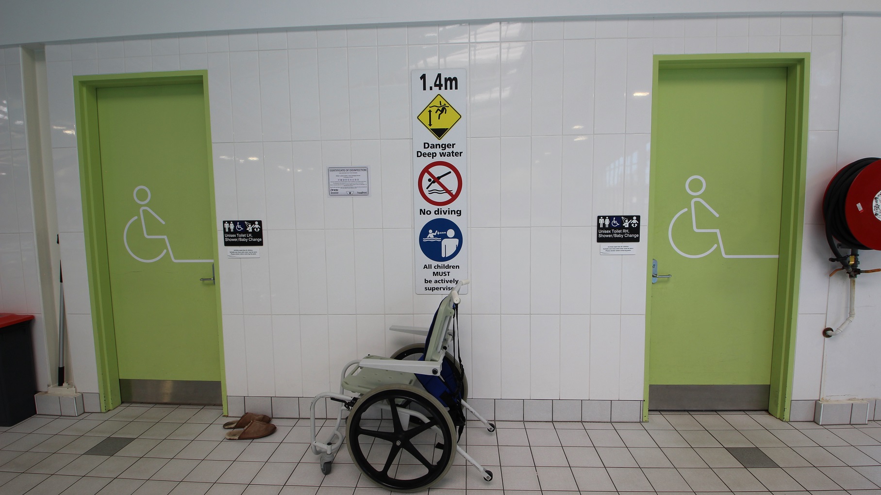 Two light green doors with wheelchair symbols are on either side of a white-tiled wall. Between the doors, a sign indicates a water depth of 1.4 metres, warning of deep water and prohibiting diving. Below the sign, a wheelchair is positioned against the wall. A pair of brown sandals is on the floor near the left door. A red fire hose reel is mounted on the wall to the right. The floor is tiled in a light grey pattern. Two light green doors with wheelchair symbols are on either side of a white-tiled wall. Between the doors, a sign indicates a water depth of 1.4 metres, warning of deep water and prohibiting diving. Below the sign, a wheelchair is positioned against the wall. A pair of brown sandals is on the floor near the left door. A red fire hose reel is mounted on the wall to the right. The floor is tiled in a light grey pattern.