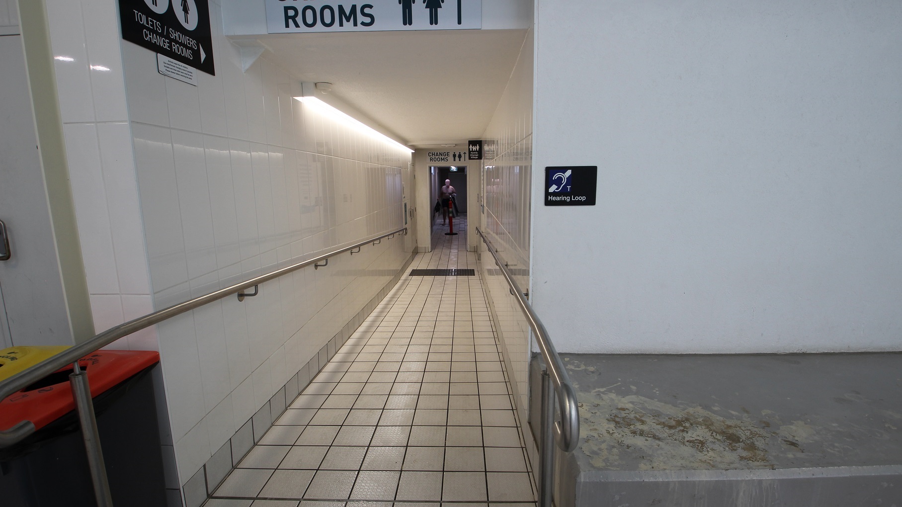 A narrow corridor with tiled walls and floor, leading to changing rooms. On the left, there's a handrail and a bin with a red lid. On the right, a sign indicates a hearing loop. Overhead, signs direct to toilets, showers, and changing rooms. At the end of the corridor, a person is visible in the distance. The ceiling is plain with fluorescent lighting. A narrow corridor with tiled walls and floor, leading to changing rooms. On the left, there's a handrail and a bin with a red lid. On the right, a sign indicates a hearing loop. Overhead, signs direct to toilets, showers, and changing rooms. At the end of the corridor, a person is visible in the distance. The ceiling is plain with fluorescent lighting.