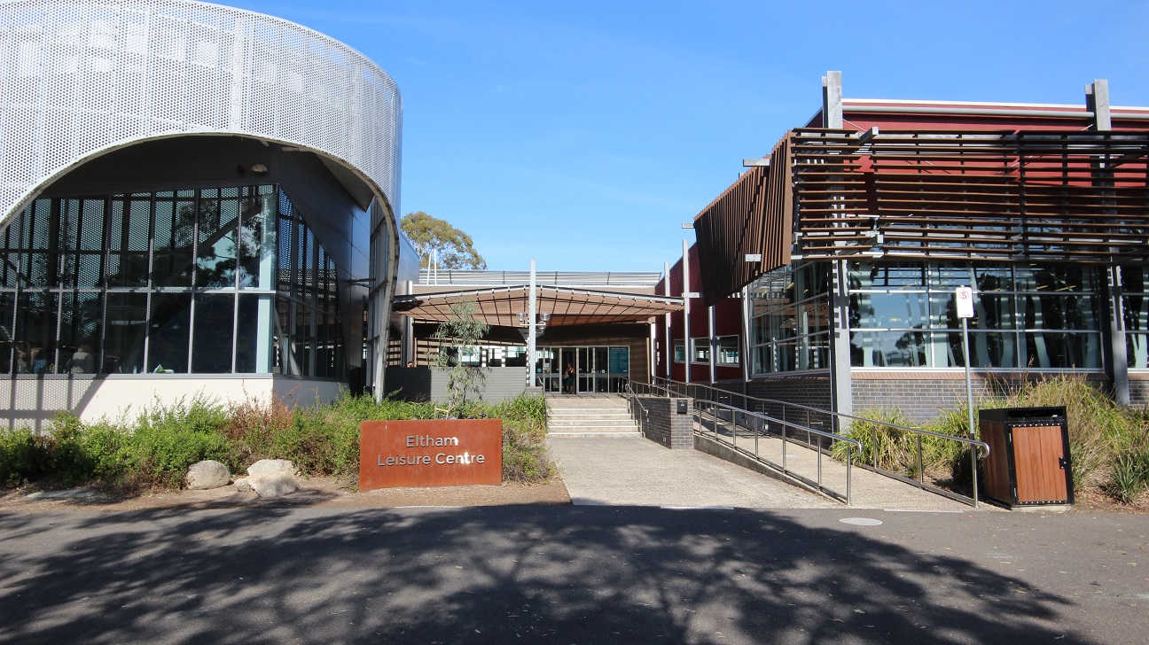 A modern building with a curved, perforated metal facade on the left and a structure with horizontal wooden slats on the right. In the centre, there is a pathway leading to the entrance, flanked by greenery. A sign reading "Eltham Leisure Centre" is visible in the foreground. The area is well-lit with clear skies, and there is a ramp and steps leading up to the entrance. A rubbish bin is positioned on the right side near the path. A modern building with a curved, perforated metal facade on the left and a structure with horizontal wooden slats on the right. In the centre, there is a pathway leading to the entrance, flanked by greenery. A sign reading "Eltham Leisure Centre" is visible in the foreground. The area is well-lit with clear skies, and there is a ramp and steps leading up to the entrance. A rubbish bin is positioned on the right side near the path.