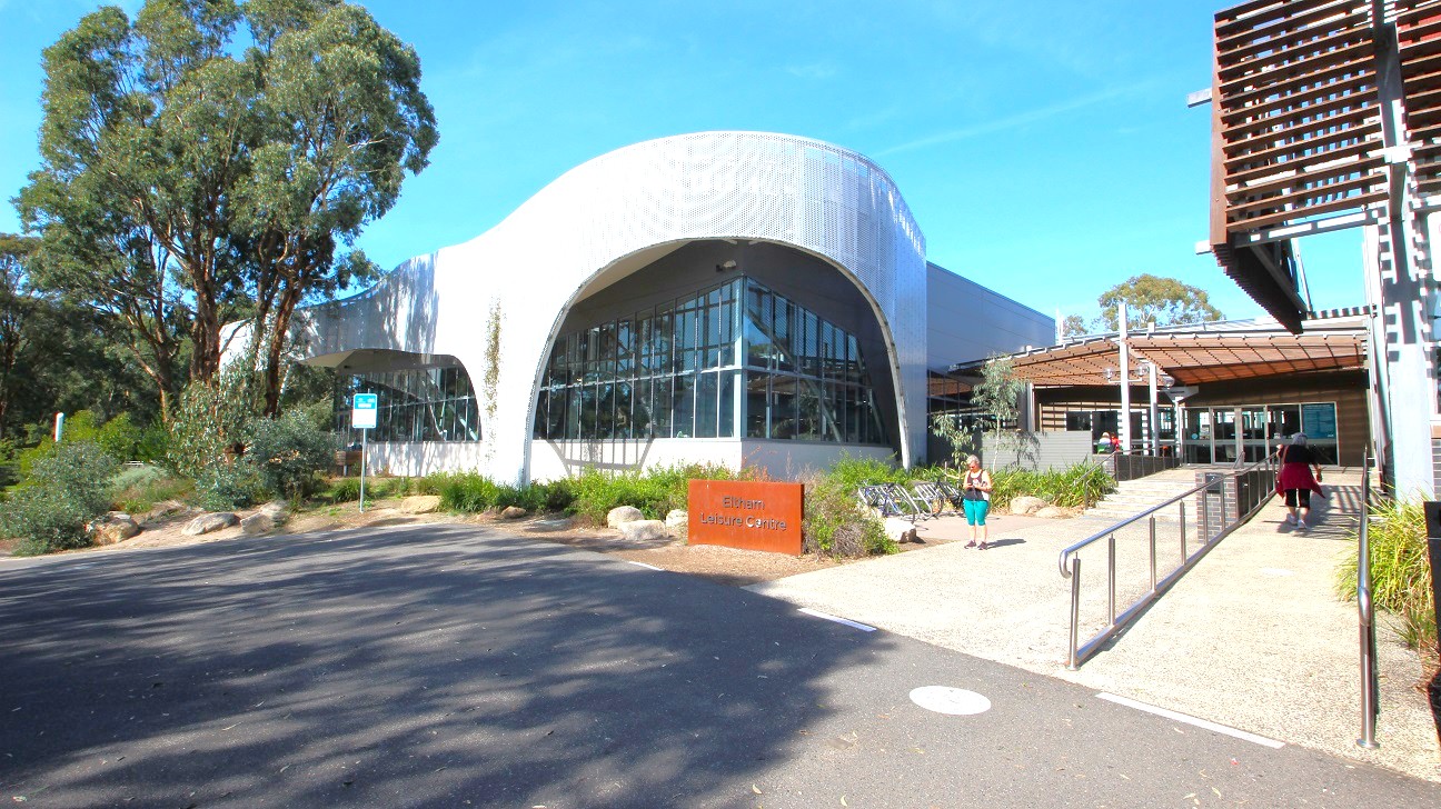 Modern building with a curved, perforated metal facade on the left and a structure with horizontal wooden slats on the right. In the centre, a pathway leads to the entrance, flanked by greenery and a sign reading "Eltham Leisure Centre". The ground is paved, and there is a ramp on the right side. The building features large glass windows, and there are trees visible in the background. Modern building with a curved, perforated metal facade on the left and a structure with horizontal wooden slats on the right. In the centre, a pathway leads to the entrance, flanked by greenery and a sign reading "Eltham Leisure Centre". The ground is paved, and there is a ramp on the right side. The building features large glass windows, and there are trees visible in the background.