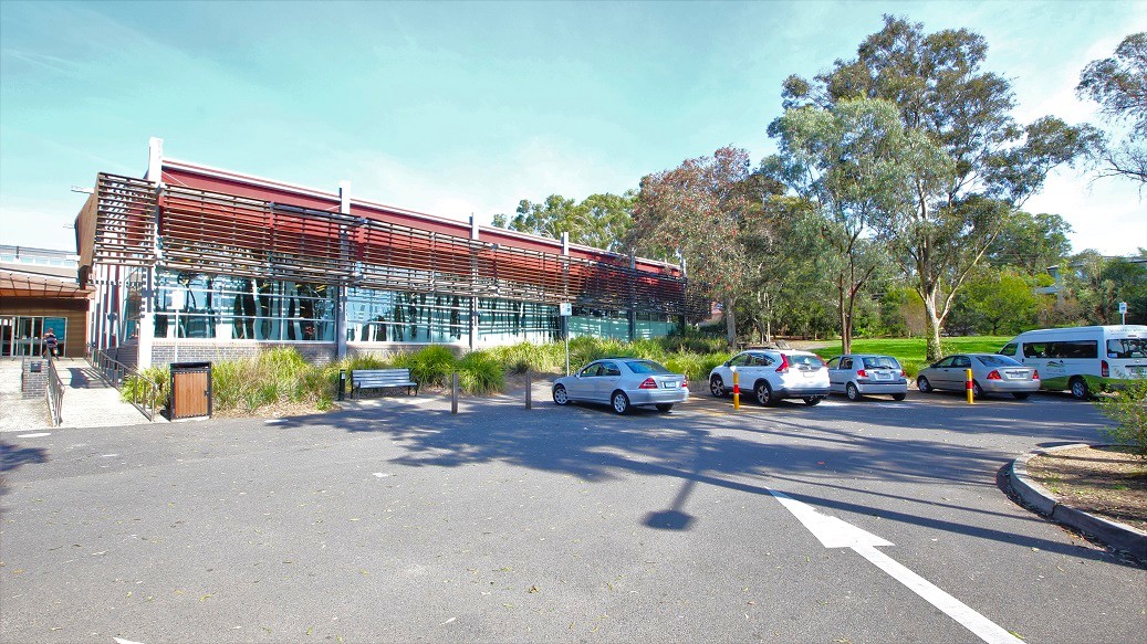 A modern building with a curved, metallic facade and large glass windows is on the left. In front, there's a sign with text and landscaped greenery. A pathway leads to the entrance on the right, where people are walking. The building is surrounded by tall trees, and there's a clear blue sky above. A railing lines the path, and the ground is a mix of pavement and gravel. A modern building with a curved, metallic facade and large glass windows is on the left. In front, there's a sign with text and landscaped greenery. A pathway leads to the entrance on the right, where people are walking. The building is surrounded by tall trees, and there's a clear blue sky above. A railing lines the path, and the ground is a mix of pavement and gravel.