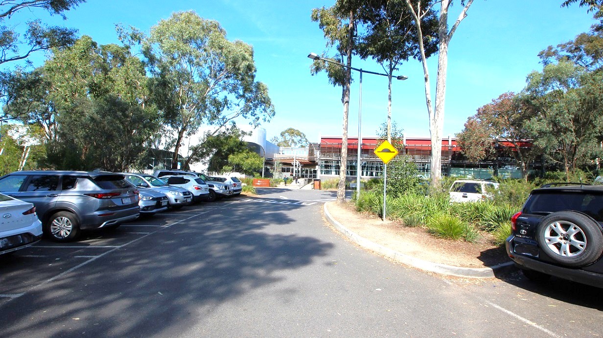 A modern building with a facade featuring horizontal slats and large glass windows is on the left. In front, there is a small parking area with several cars and a minibus. The ground is paved with asphalt, and there are some bollards near the parking spaces. On the right, there are tall trees and green grass, suggesting a park or garden area. The sky is clear, and the scene is well-lit, indicating daytime. A modern building with a facade featuring horizontal slats and large glass windows is on the left. In front, there is a small parking area with several cars and a minibus. The ground is paved with asphalt, and there are some bollards near the parking spaces. On the right, there are tall trees and green grass, suggesting a park or garden area. The sky is clear, and the scene is well-lit, indicating daytime.