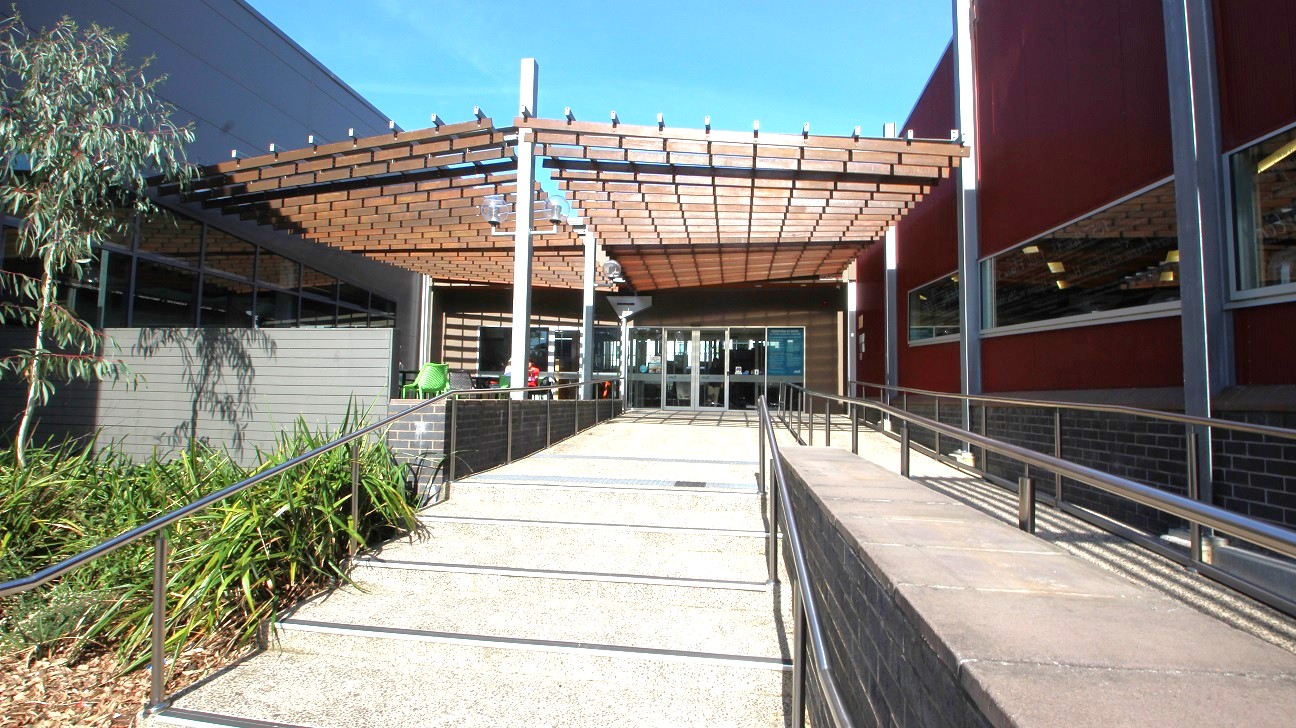 A car park with several vehicles lined up on the left side, including a grey SUV and white cars. On the right, a yellow road sign is visible near some greenery. In the background, a building with a red roof and large windows is partially obscured by trees. The road curves slightly, and tall trees are scattered throughout the area. The sky is clear and blue. A car park with several vehicles lined up on the left side, including a grey SUV and white cars. On the right, a yellow road sign is visible near some greenery. In the background, a building with a red roof and large windows is partially obscured by trees. The road curves slightly, and tall trees are scattered throughout the area. The sky is clear and blue.