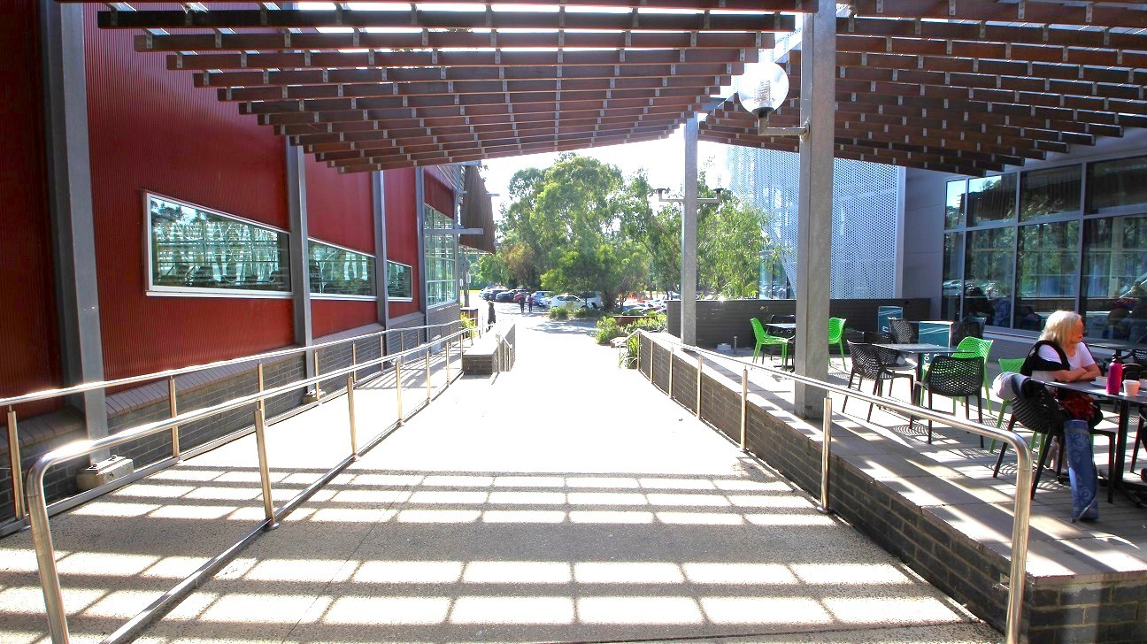 A pathway with metal railings runs through the centre, leading to an outdoor area with trees and parked cars in the background. On the left, a red building with long horizontal windows. On the right, a seating area with tables and chairs, where a person is sitting. Above, a grid-like structure provides partial shade, casting shadows on the ground. A pathway with metal railings runs through the centre, leading to an outdoor area with trees and parked cars in the background. On the left, a red building with long horizontal windows. On the right, a seating area with tables and chairs, where a person is sitting. Above, a grid-like structure provides partial shade, casting shadows on the ground.