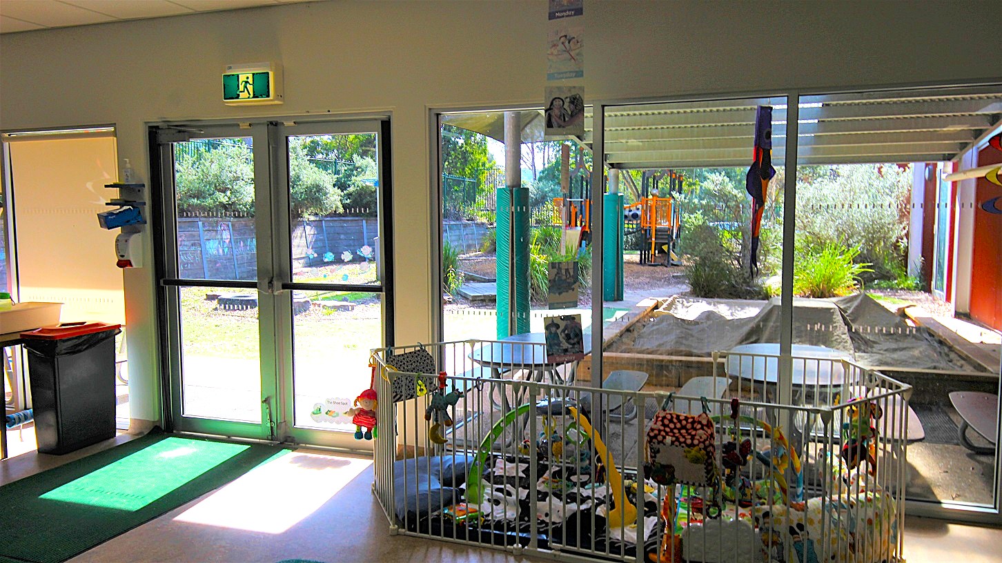 White fence at the entrance Child Care Centre at Eltham Leisure Centre. Beyond the fence are toys, costumes, kids' decorations, kids' drawings and paintings, and a soft green mat to protect children during playing. White fence at the entrance Child Care Centre at Eltham Leisure Centre. Beyond the fence are toys, costumes, kids' decorations, kids' drawings and paintings, and a soft green mat to protect children during playing.