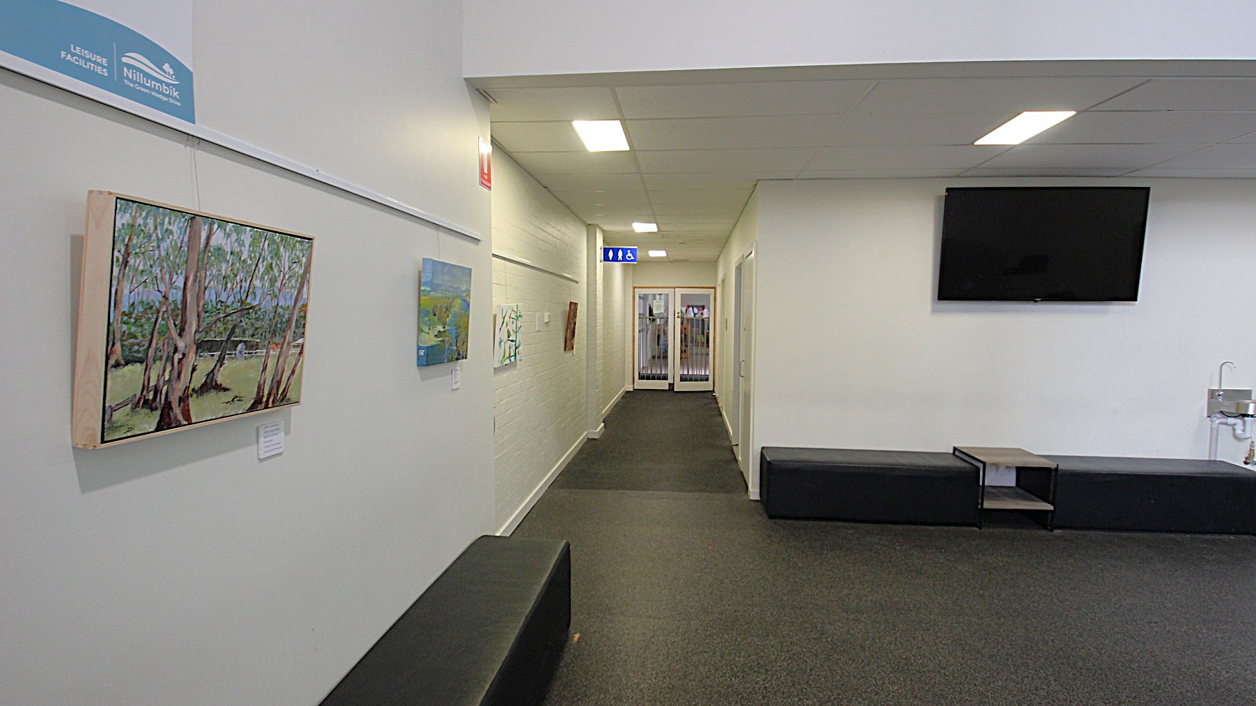 A small space with TV, paintings, and black sofa chairs in the white corridor at the entrance of Child Care at Eltham Leisure Centre. Underneath the TV is a faucet and between the sofa chairs is a magazine table. A small space with TV, paintings, and black sofa chairs in the white corridor at the entrance of Child Care at Eltham Leisure Centre. Underneath the TV is a faucet and between the sofa chairs is a magazine table.