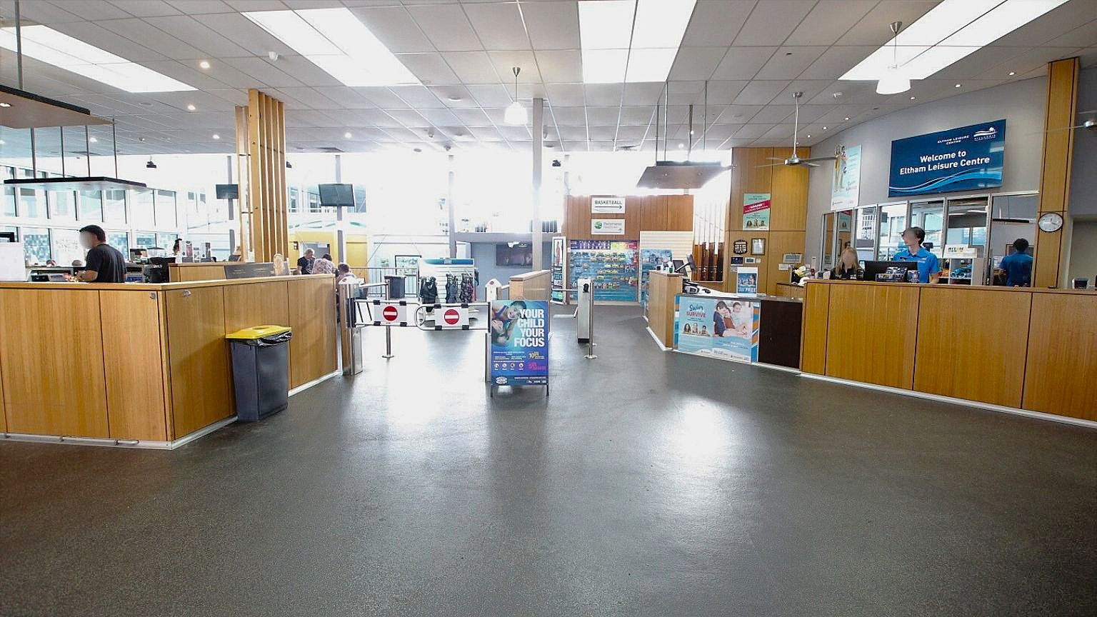 A spacious interior with a dark floor and a high ceiling featuring large rectangular lights. On the left, a wooden counter with a yellow-lidded bin and a person behind it. In the centre, a sign and a barrier with "no entry" symbols. On the right, another wooden counter with two people behind it, and a digital sign above reading "Welcome to Eltham Leisure Centre." The background has glass panels and more signage. A spacious interior with a dark floor and a high ceiling featuring large rectangular lights. On the left, a wooden counter with a yellow-lidded bin and a person behind it. In the centre, a sign and a barrier with "no entry" symbols. On the right, another wooden counter with two people behind it, and a digital sign above reading "Welcome to Eltham Leisure Centre." The background has glass panels and more signage.