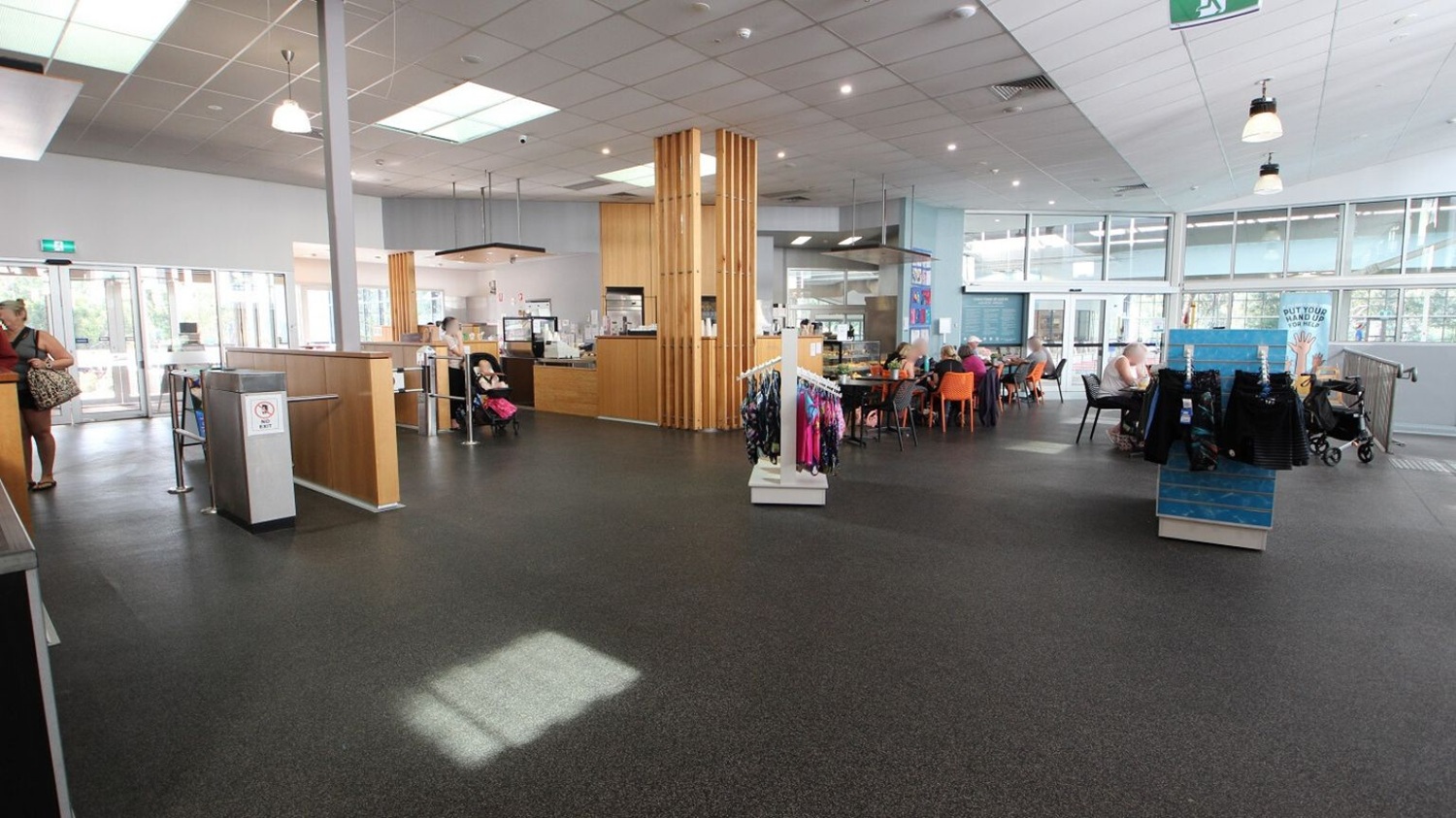 A spacious indoor area with a dark floor and a ceiling featuring recessed lighting and skylights. On the left, a woman stands near a counter with a glass entrance behind her. In the centre, a wooden column and a small café area with people seated. On the right, a display rack with clothes and a group of people sitting at tables with orange chairs. Large windows line the background, allowing natural light to fill the space. A spacious indoor area with a dark floor and a ceiling featuring recessed lighting and skylights. On the left, a woman stands near a counter with a glass entrance behind her. In the centre, a wooden column and a small café area with people seated. On the right, a display rack with clothes and a group of people sitting at tables with orange chairs. Large windows line the background, allowing natural light to fill the space.