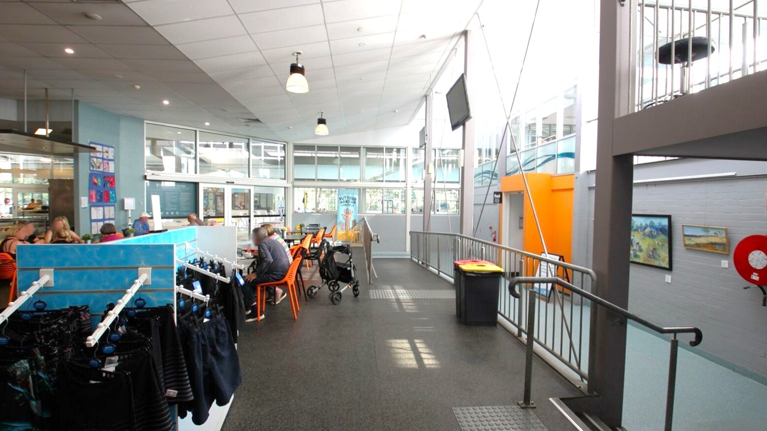 A spacious indoor area with a high ceiling and large windows on the right, allowing natural light to flood in. On the left, there are people seated at tables with orange chairs, and a blue partition with hanging items. In the middle, a pram is parked near the tables. The floor is dark and textured, with a ramp and railing leading to a higher level. On the right wall, there are paintings and a red fire hose reel. The ceiling has several hanging lights. A spacious indoor area with a high ceiling and large windows on the right, allowing natural light to flood in. On the left, there are people seated at tables with orange chairs, and a blue partition with hanging items. In the middle, a pram is parked near the tables. The floor is dark and textured, with a ramp and railing leading to a higher level. On the right wall, there are paintings and a red fire hose reel. The ceiling has several hanging lights.