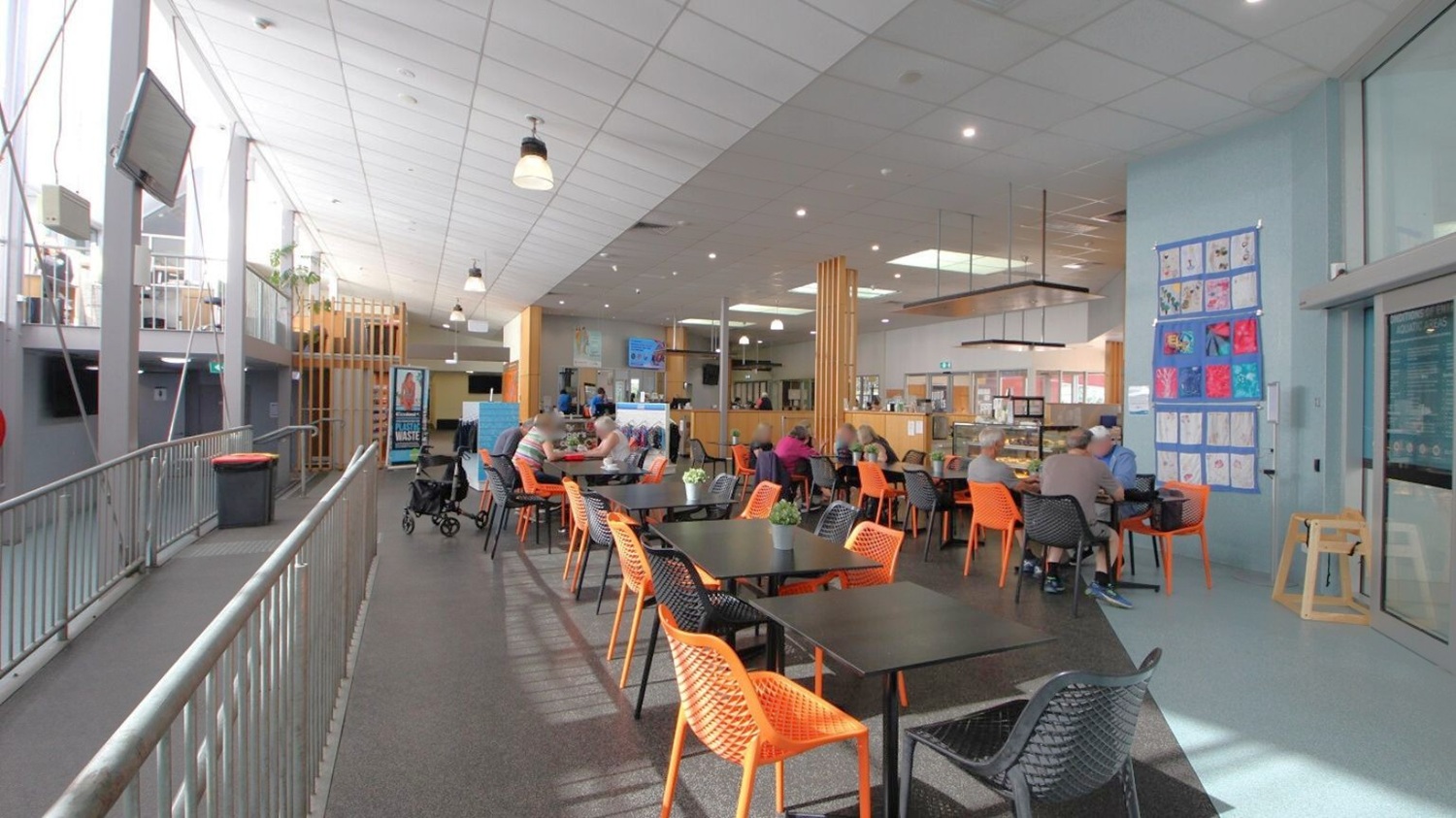A spacious indoor café area with a high ceiling and modern design. On the left, a metal railing lines a walkway with a mounted TV screen above. The floor is a mix of dark and light tiles. In the centre, there are black tables with orange and black woven chairs. People are seated, engaged in conversation. The right side features a light blue wall adorned with colourful artwork. The café counter is visible in the background, with hanging lights and a menu display. A spacious indoor café area with a high ceiling and modern design. On the left, a metal railing lines a walkway with a mounted TV screen above. The floor is a mix of dark and light tiles. In the centre, there are black tables with orange and black woven chairs. People are seated, engaged in conversation. The right side features a light blue wall adorned with colourful artwork. The café counter is visible in the background, with hanging lights and a menu display.