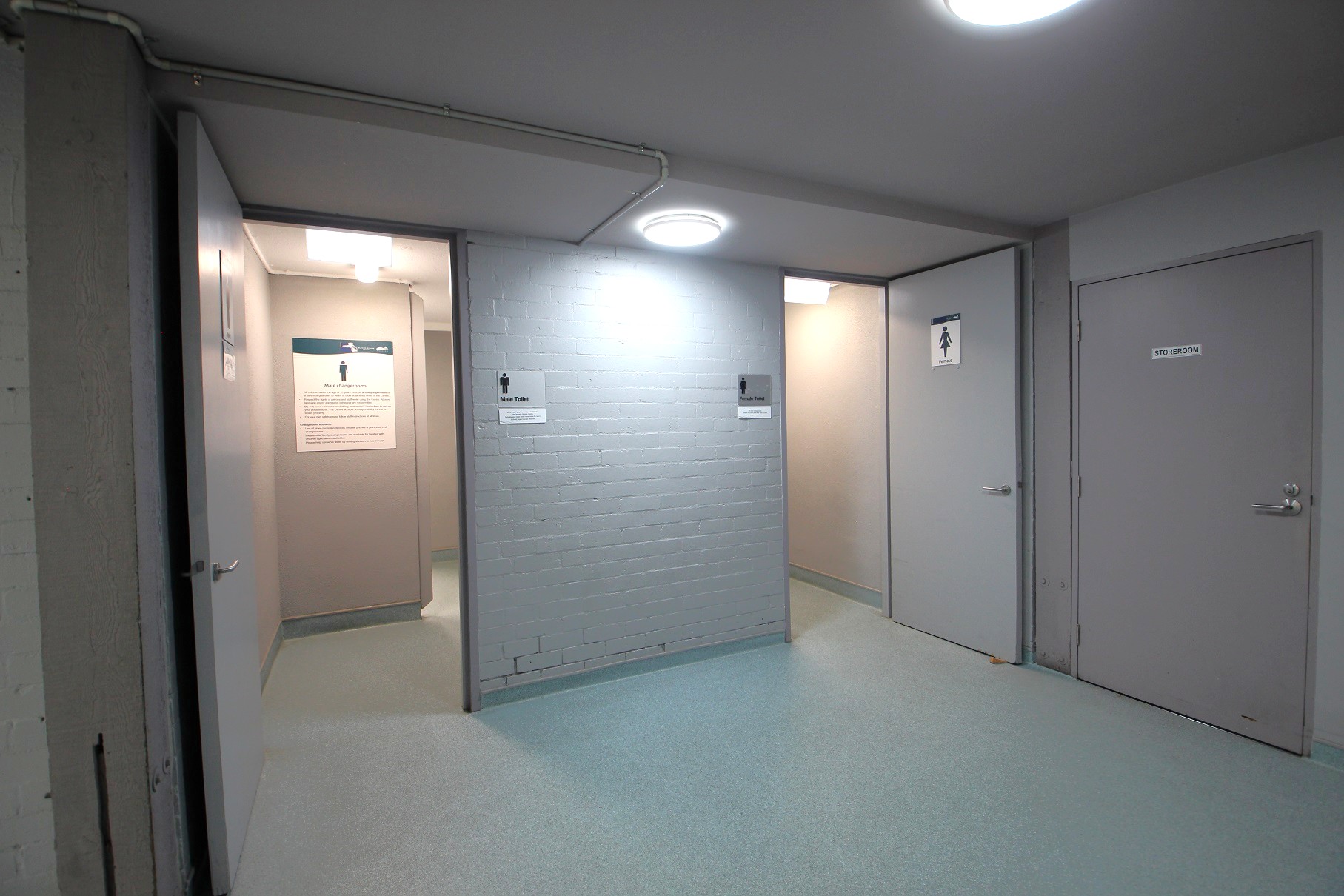 A corridor with three doors. On the left, a door is open, leading to a room with a sign indicating a male toilet. In the centre, a wall with a sign for male toilets. On the right, two closed doors; one marked with a female toilet sign and the other labelled "Storeroom". The floor is light-coloured, and the ceiling has round, recessed lights. A corridor with three doors. On the left, a door is open, leading to a room with a sign indicating a male toilet. In the centre, a wall with a sign for male toilets. On the right, two closed doors; one marked with a female toilet sign and the other labelled "Storeroom". The floor is light-coloured, and the ceiling has round, recessed lights.