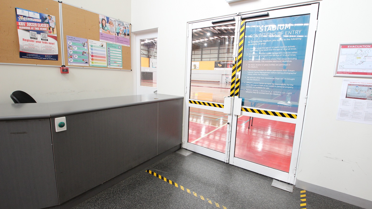 Adjacent to the dark grey reception desk at the entrance of Eltham Leisure Centre is the glass door with yellow and black safety tape. On the right side of the door is an evacuation map. Adjacent to the dark grey reception desk at the entrance of Eltham Leisure Centre is the glass door with yellow and black safety tape. On the right side of the door is an evacuation map.