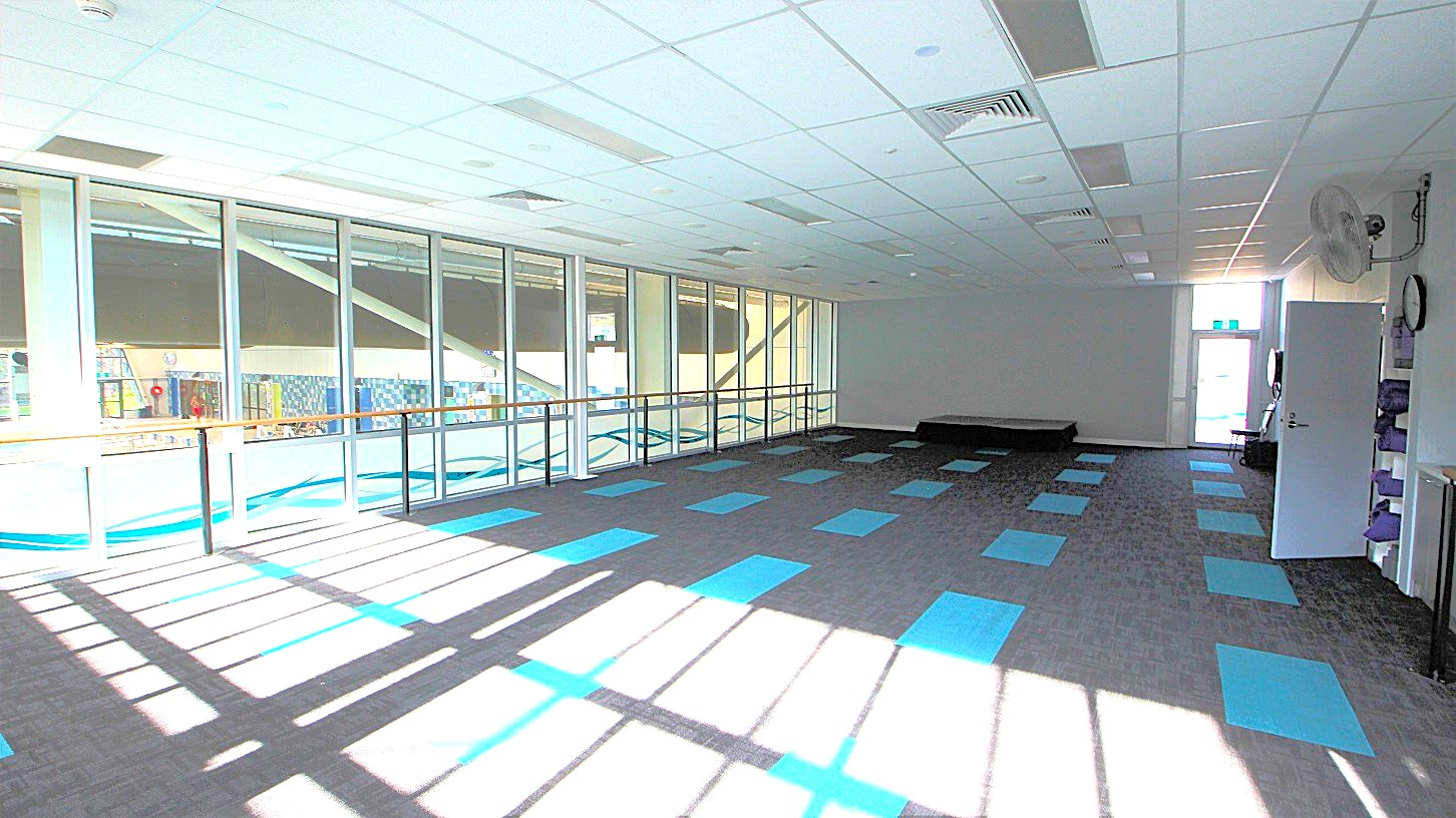A well-ventilated yoga studio with glass walls, grey and blue rectangular carpet tiles, and wooden platforms for handheld during yoga practice. The front wall is painted grey while the ceilings are purely white. A well-ventilated yoga studio with glass walls, grey and blue rectangular carpet tiles, and wooden platforms for handheld during yoga practice. The front wall is painted grey while the ceilings are purely white.