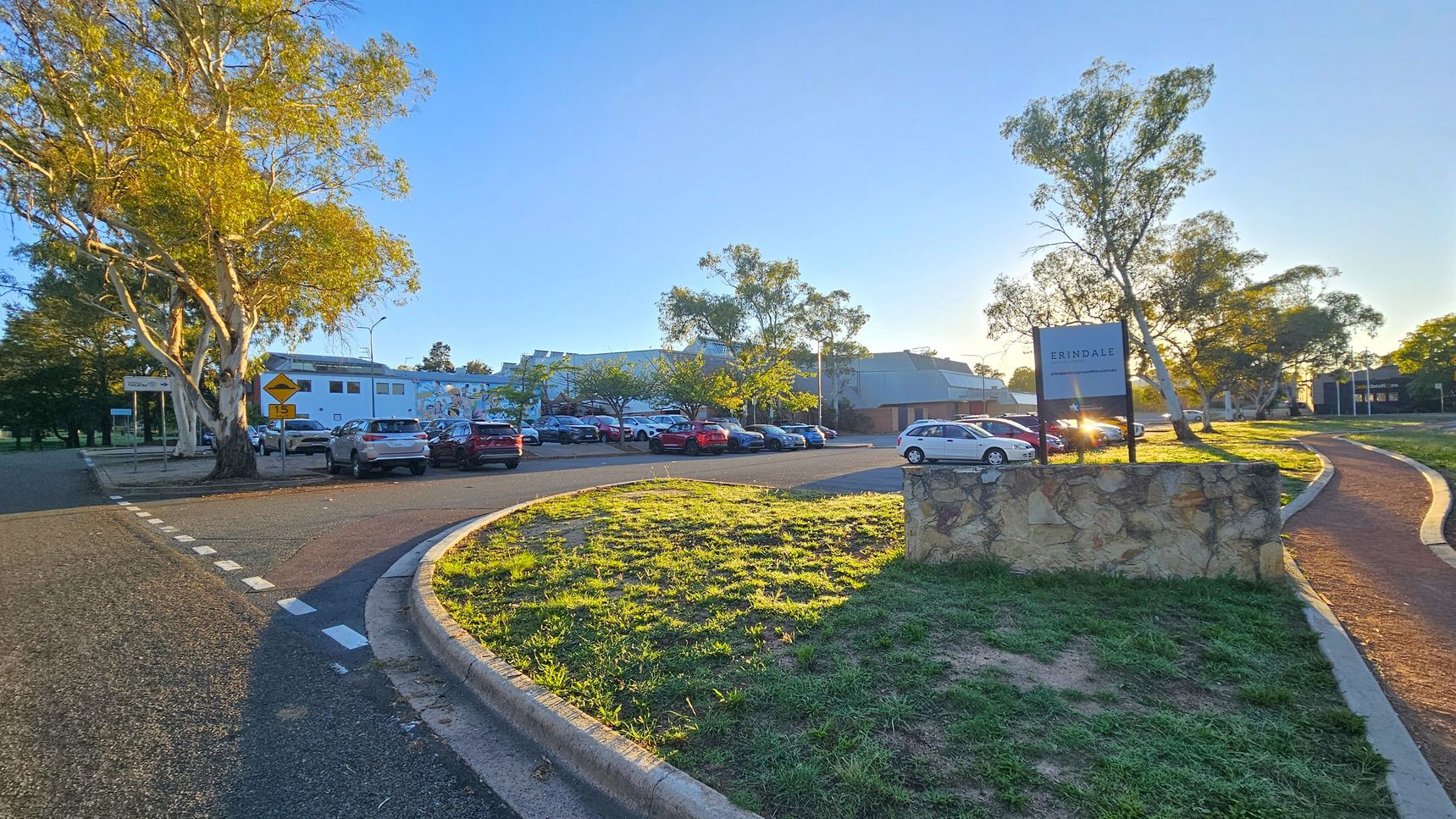 A road curves from the left foreground to the centre, bordered by a grassy area. On the right, a stone sign reads "ERINDALE". Several cars are parked in a lot in the middle distance, with a building behind them. Tall trees with light green foliage are scattered throughout the scene. The sun is low, casting long shadows and illuminating the area with a warm glow. A pedestrian path runs parallel to the road on the right.