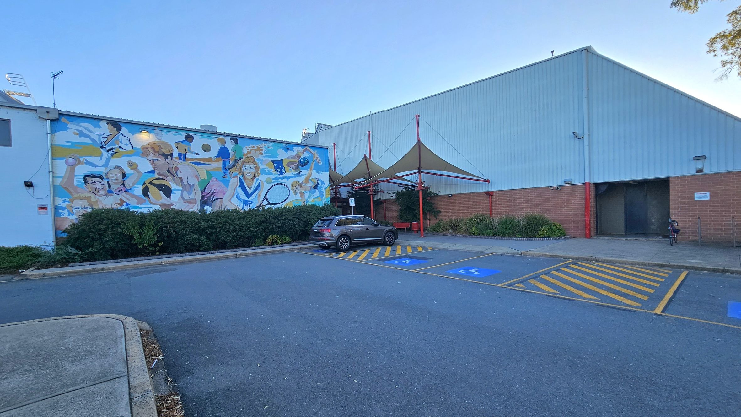 A car park with designated disabled parking spaces marked in blue and yellow lines. On the left, a large mural on a building wall depicts various sports activities. In the centre, a parked car is near the mural. To the right, a building with a corrugated metal roof and brick walls features a shaded area with triangular canopies supported by red poles. Bushes and small plants line the building's base. The ground is asphalt, and the area is well-maintained.