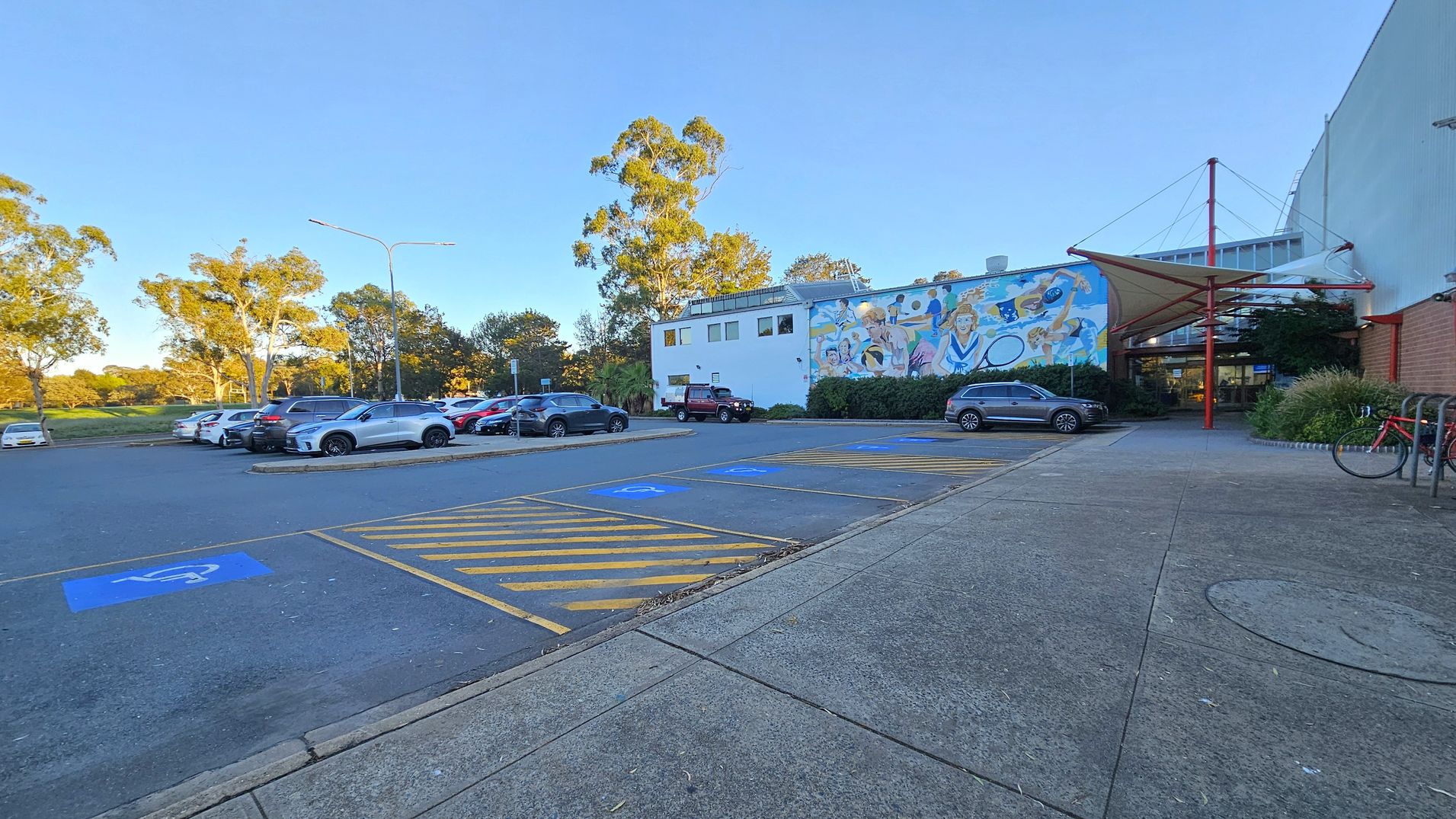 A car park with several vehicles on the left, including a silver car in the foreground. The ground has marked disabled parking spaces with blue and yellow lines. In the centre, a building with a colourful mural depicting people engaged in various activities. On the right, a covered entrance with red structural elements and a bicycle parked nearby. Trees and greenery are visible in the background. The pavement is concrete.