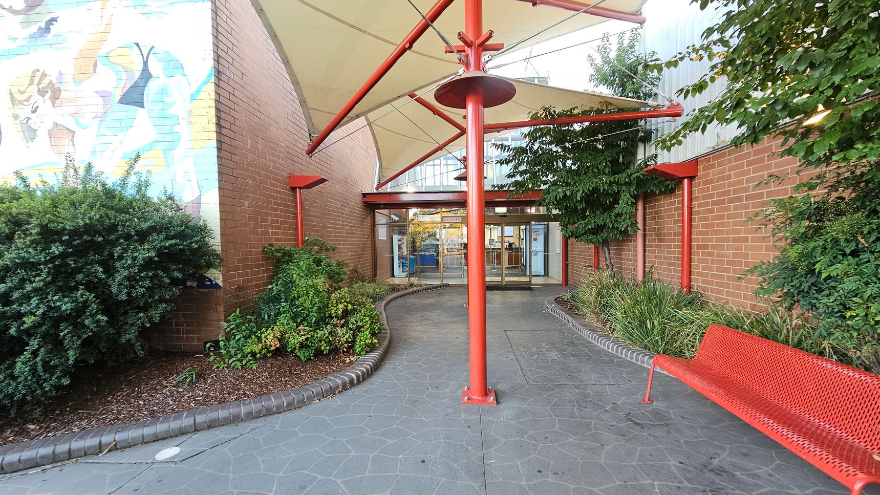 A pathway leads to a glass entrance with a red metal frame. On the left, a brick wall features a colourful mural partially visible above dense shrubs. On the right, a red metal bench sits beside a brick wall with lush greenery. A large red pole supports a cream-coloured canopy overhead. The ground is paved with grey stones, and the entrance is flanked by glass doors.