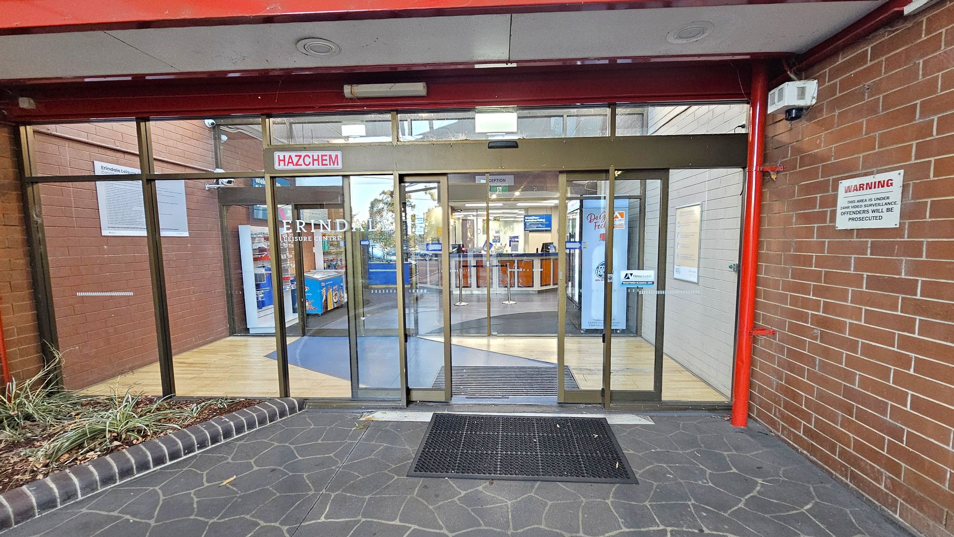 Entrance to a building with glass sliding doors. Above the doors, a "HAZCHEM" sign is visible. The floor outside is paved with dark stone tiles, and a black mat is placed in front of the entrance. Inside, a wooden floor is visible, leading to a reception area. The walls are brick, with a warning sign on the right indicating surveillance. A vending machine is visible inside on the left. The ceiling is white with red beams.