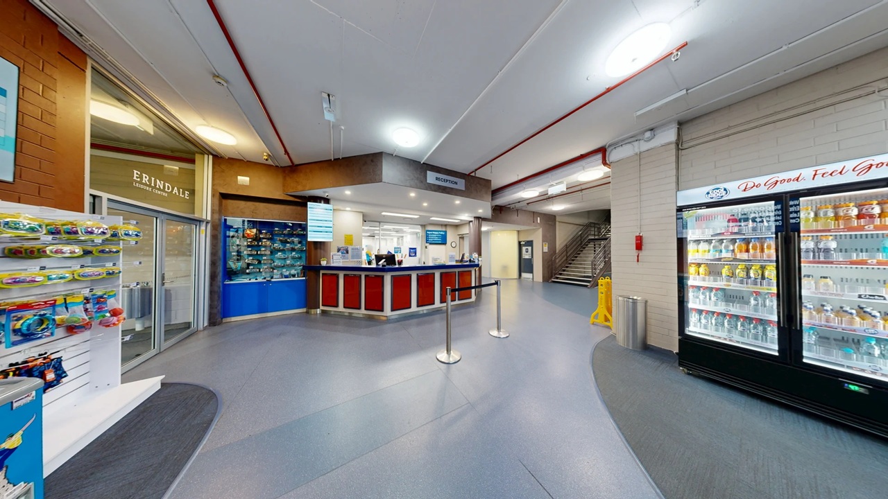 Reception area with a counter in the centre, featuring red and blue panels. To the left, a display rack with various items and a glass entrance labelled "Erindale". On the right, a large refrigerated display filled with drinks. A staircase is visible in the background, and a yellow caution sign is on the floor. The ceiling has exposed pipes and fluorescent lighting. The floor is covered with grey and blue tiles.