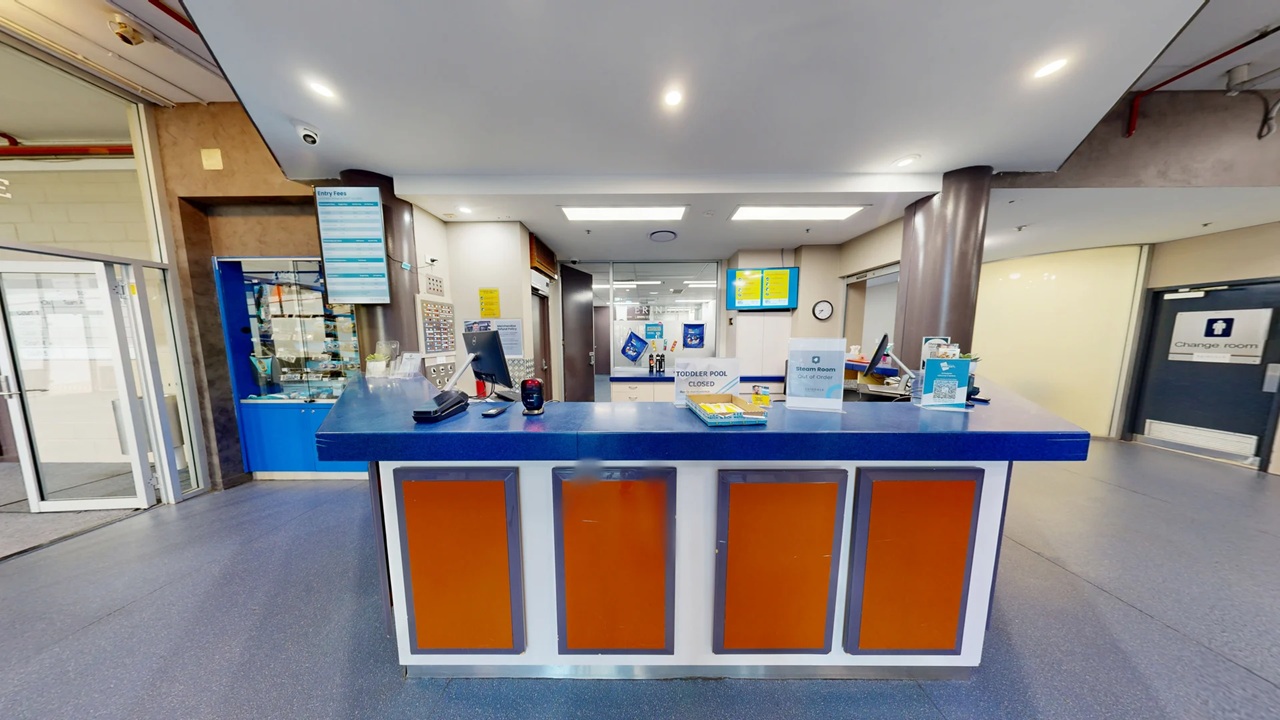 A reception area with a blue and orange counter in the centre. Behind the counter are computer monitors and various informational signs. To the left, there's a glass display case and a doorway with glass doors. On the right, a wall with a door featuring a sign. The ceiling is white with recessed lighting, and the floor is grey. In the background, there are more signs and a clock on the wall.