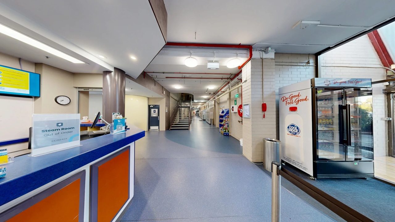 A hallway with a blue and orange reception desk on the left, featuring a sign for a steam room. A clock and a digital screen are mounted on the wall behind the desk. On the right, there's a glass-fronted refrigerator with a "Feel Good" sign. The corridor extends towards the back, with a staircase leading upwards. The floor is grey, and the ceiling has exposed pipes and lighting fixtures. A fire extinguisher and hose reel are mounted on the right wall.