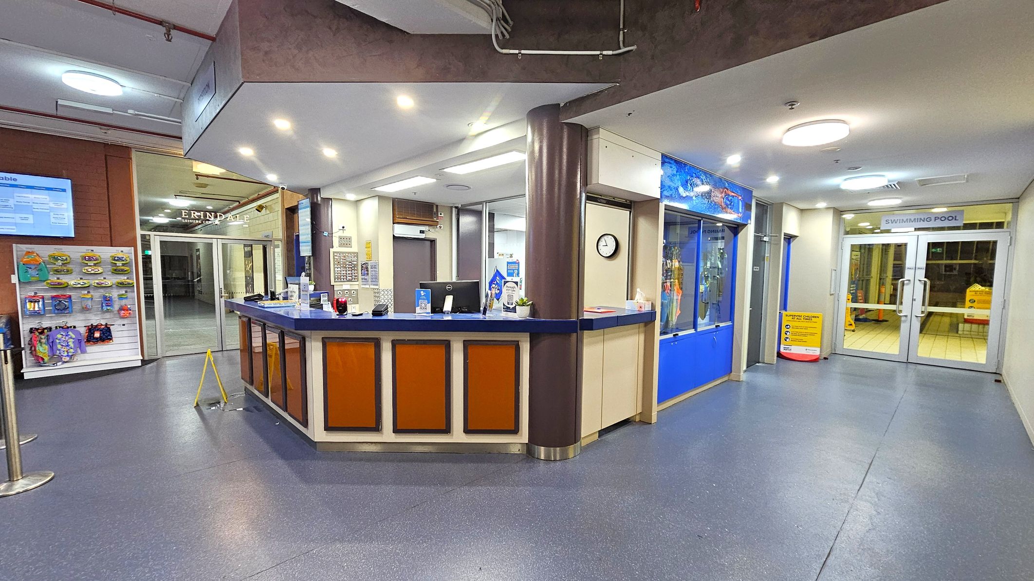 Reception area with a curved desk in the centre, featuring a computer, hand sanitiser, and a clock on the wall. To the left, a display rack with swimwear and accessories. Behind the desk, a glass door with "Erindale Leisure Centre" signage. On the right, a corridor leads to double doors marked "Swimming Pool". The floor is blue, and the ceiling has circular lights.