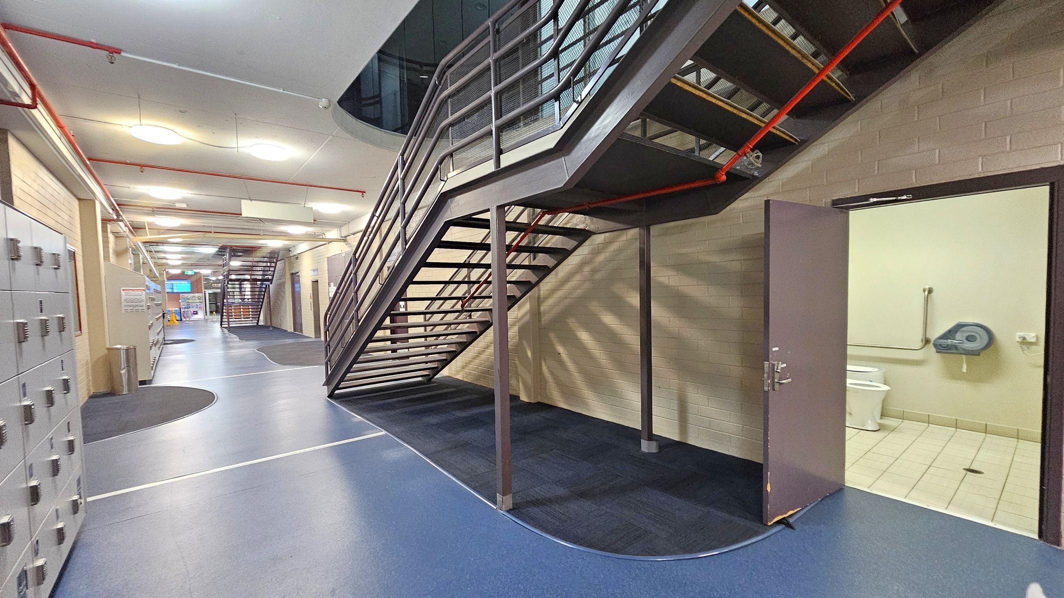 A corridor with a blue and grey floor and a staircase with metal railings leading upwards. On the right, an open door reveals a bathroom with a toilet and a paper dispenser. Lockers line the left side of the corridor. The ceiling features circular lights and exposed red pipes.