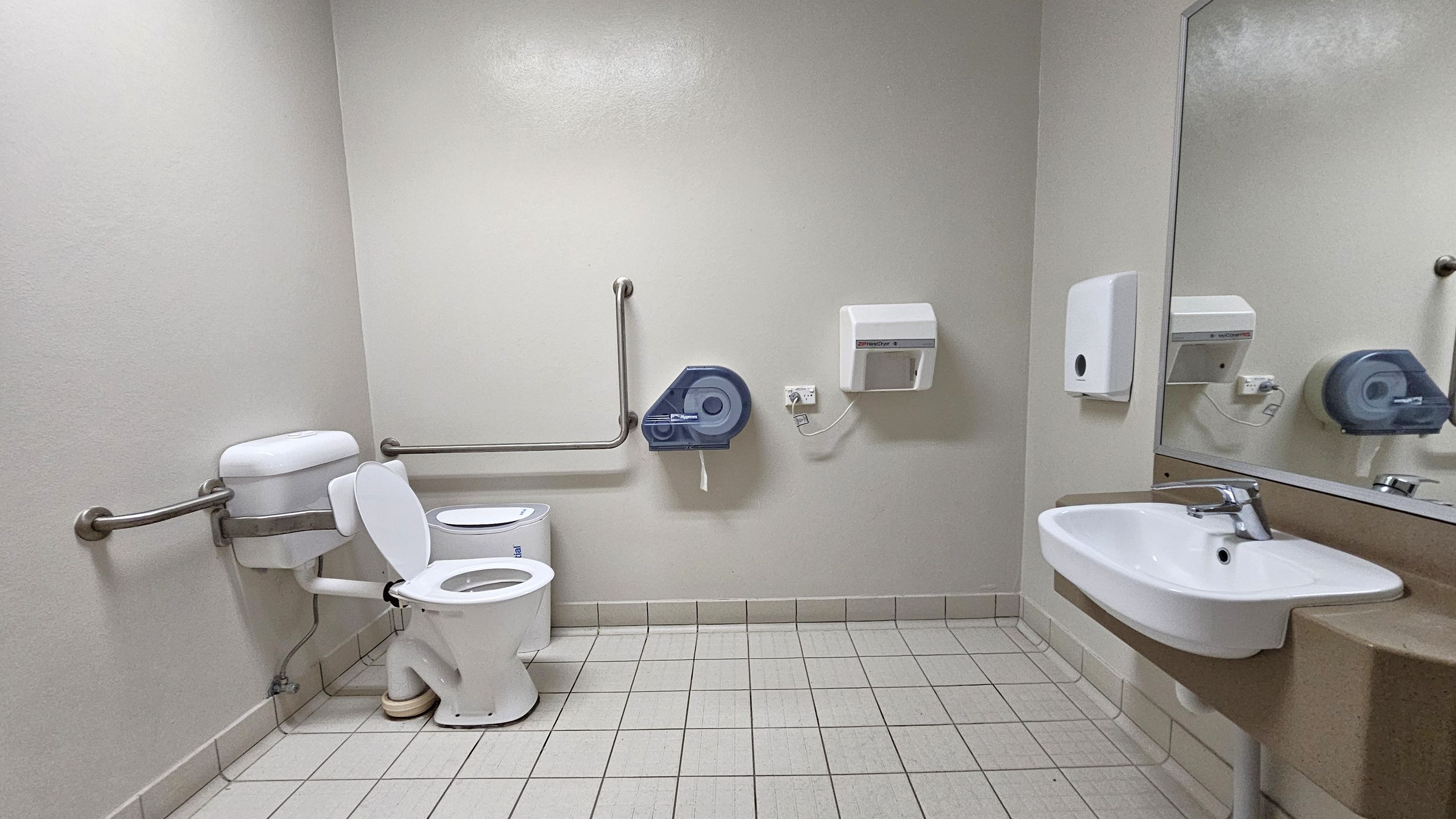 A public restroom with a white toilet on the left, equipped with metal grab bars. A large blue toilet paper dispenser is mounted on the wall beside it. To the right, a white sink with a silver tap is set into a brown countertop, with a large mirror above. A soap dispenser and a paper towel dispenser are mounted on the wall near the sink. The floor is covered with light grey tiles, and the walls are painted a light grey.