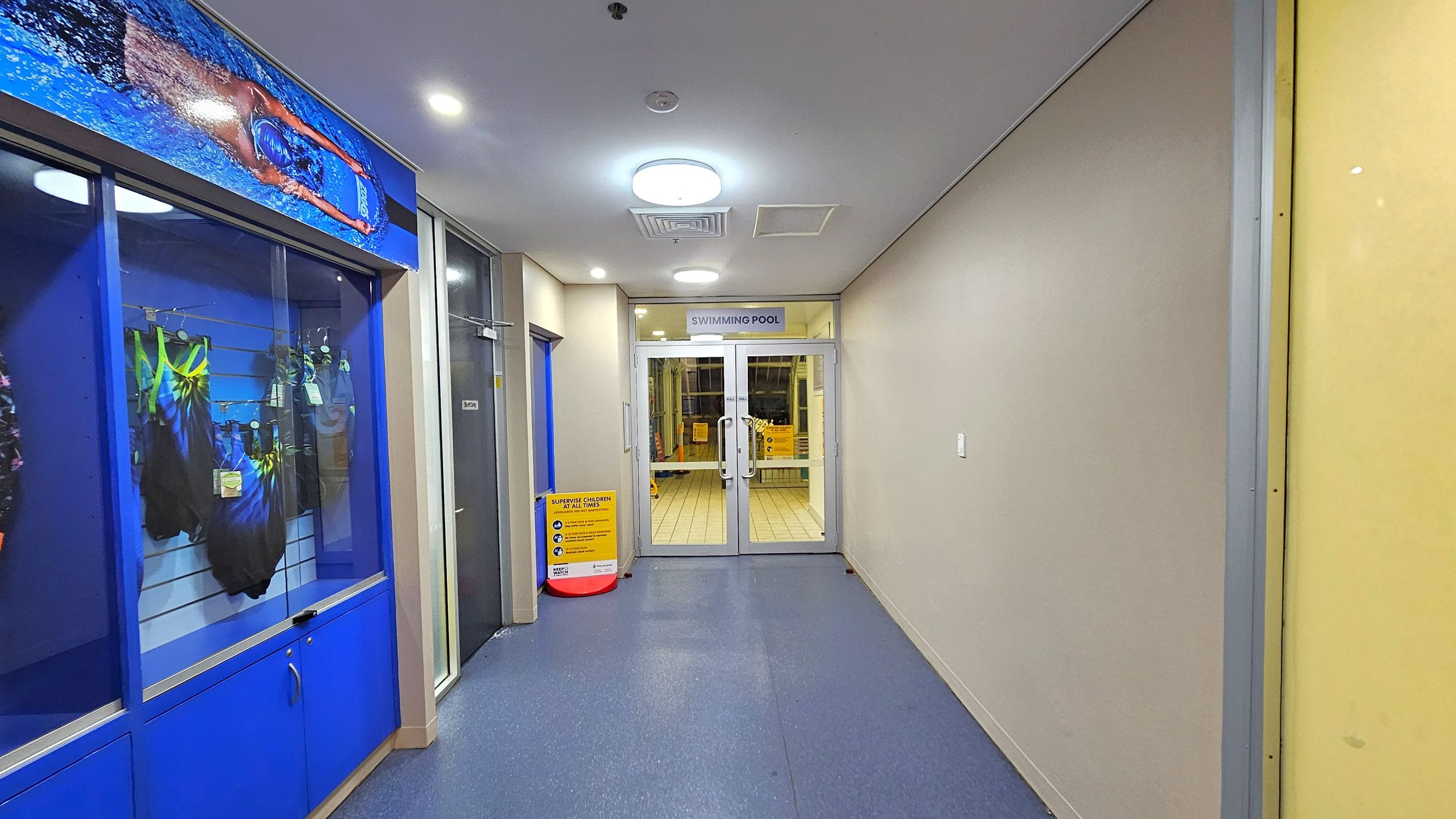 A corridor with a blue floor and a white ceiling with circular lights. On the left, there is a display case with swimming costumes and a large image of a swimmer above it. On the right, a plain wall. At the end, glass doors with a sign above reading "SWIMMING POOL". A yellow caution sign is visible near the doors.