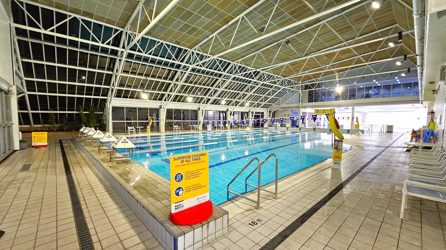 An indoor swimming pool with a tiled deck. On the left, there are "No Diving" signs on the pool's edge. A yellow sign warns to supervise children. The pool has lane dividers and a metal ladder. The right side has a row of white chairs and pool equipment. The ceiling is high with a metal framework and large windows, allowing natural light. The floor is tiled, and the pool is marked as 1.8 metres deep.