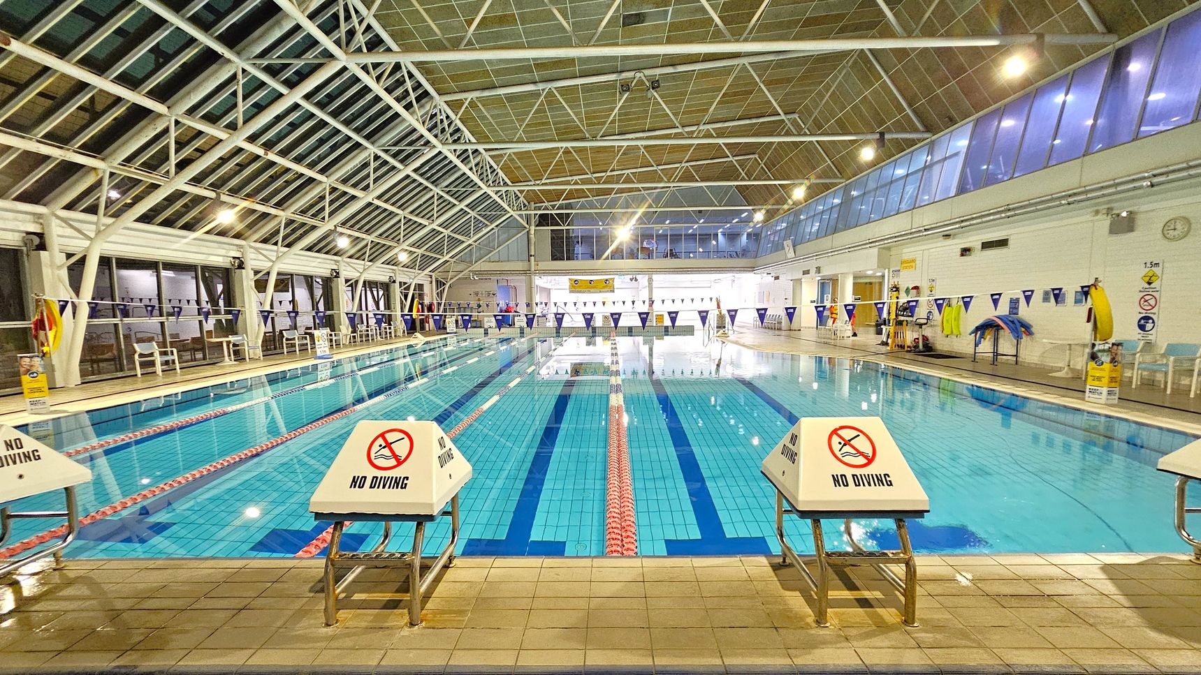 An indoor swimming pool with clear blue water, divided into lanes by red and white ropes. "No Diving" signs are visible on the starting blocks at the pool's edge. The left side has a row of white chairs and large windows with a metal framework. The right side features a white wall with safety equipment and a clock. The ceiling is high with a grid of metal beams and lights, and the floor is tiled. Blue and white triangular flags hang above the pool.