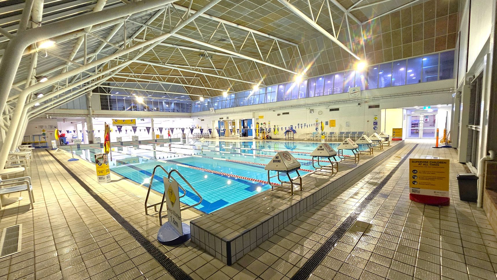 An indoor swimming pool with multiple lanes, surrounded by tiled flooring. On the left, there are benches and a lifeguard station. The right side features a sign warning to supervise children. The pool is bordered by "No Diving" signs. The ceiling is high with a metal framework and large windows on the upper walls, allowing natural light. The far end has doors and additional signage.