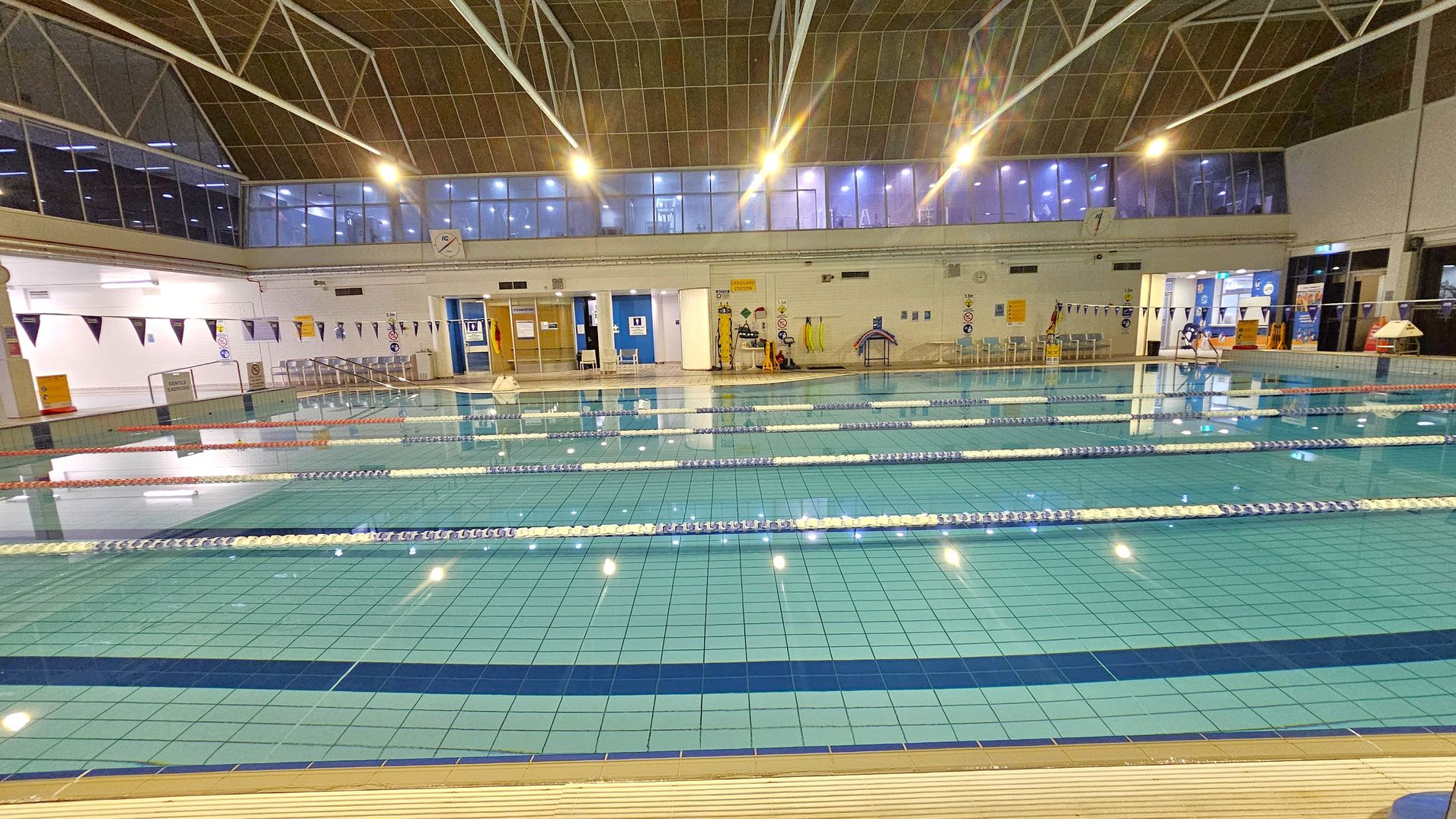 An indoor swimming pool with multiple lanes separated by floating lane dividers. The pool area is surrounded by a tiled deck. On the far side, there are doors and windows, with some signage and equipment like lifebuoys and kickboards. Above, large windows and a high ceiling with exposed beams are visible. The lighting is bright, with several overhead lights illuminating the space.