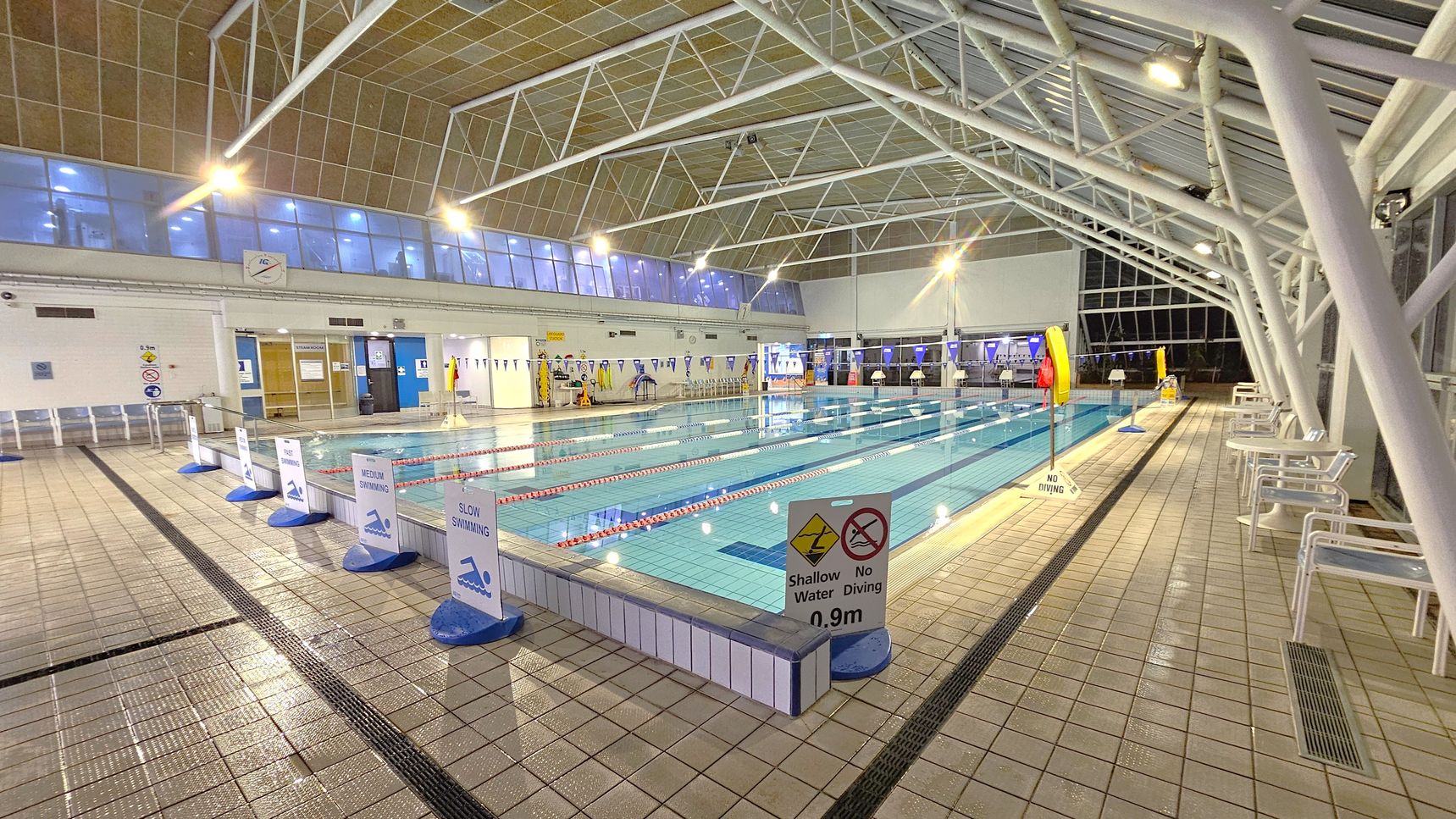 Indoor swimming pool with lanes marked by red and white ropes. Signs indicate "Fast", "Medium", and "Slow" swimming lanes. The pool is surrounded by a tiled deck with white chairs on the right. Overhead, a high ceiling with metal beams and large windows allows natural light. Signs warn of shallow water and no diving. The left side has changing rooms and additional signage. The pool area is well-lit with overhead lights.