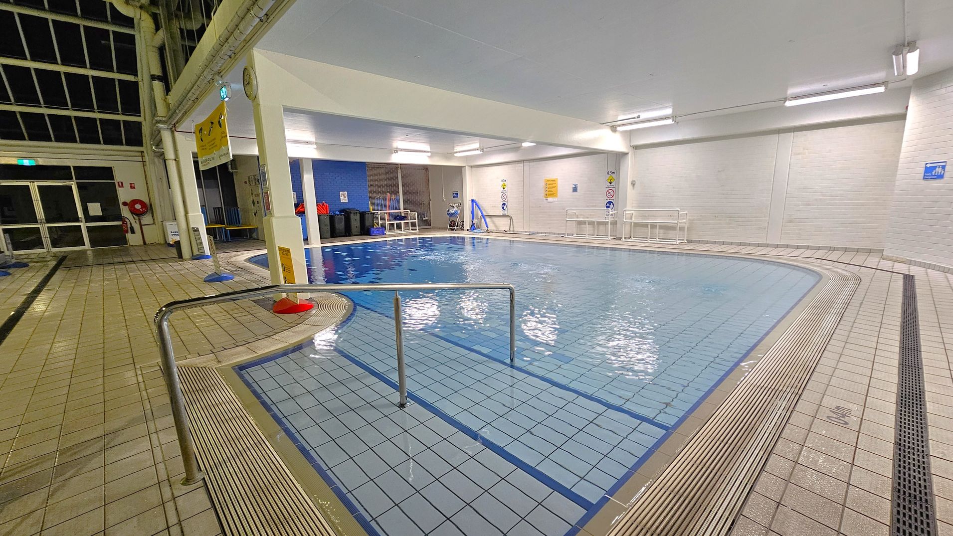Indoor swimming pool with light blue tiles and a metal handrail leading into the water. The pool is surrounded by a tiled deck. On the left, there are large windows and a door, with a fire extinguisher and a red hose reel on the wall. The right side has white brick walls with safety signs. Overhead, fluorescent lights illuminate the area. In the background, there are benches and equipment against a blue wall.