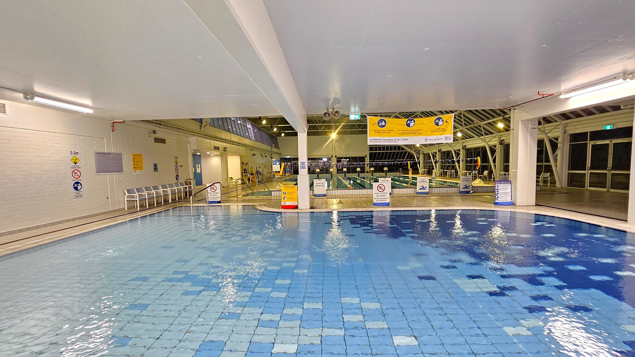 Indoor swimming pool with a tiled floor in varying shades of blue. On the left, a white brick wall with safety signs and a row of white chairs. In the centre, a yellow banner with pool rules hangs above the water. The pool deck has several signs and barriers. On the right, large windows and doors with metal frames. The ceiling is white with exposed beams and lighting fixtures.
