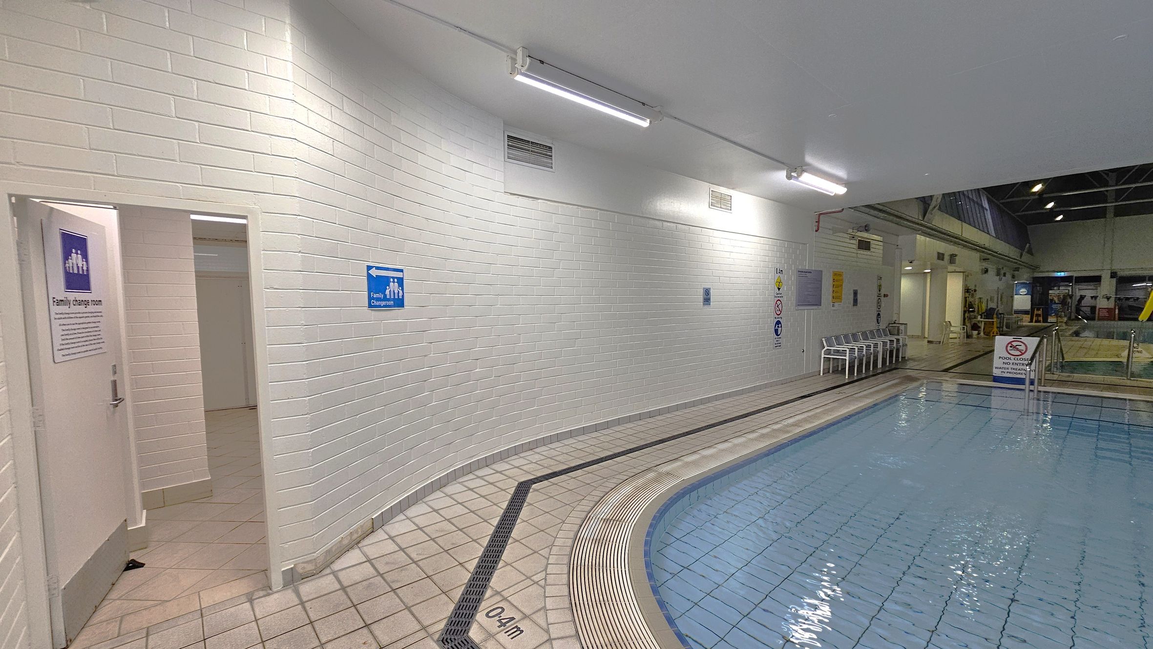 A swimming pool area with a tiled floor and a shallow pool marked "0.4m" on the right. The pool is bordered by a non-slip surface. On the left, a white brick wall curves around, featuring a door labelled "Family change room" and a blue sign pointing towards it. Several white chairs are lined up along the wall near the pool. Overhead, fluorescent lights illuminate the space, and the ceiling has exposed beams. In the background, more pool facilities and equipment are visible.