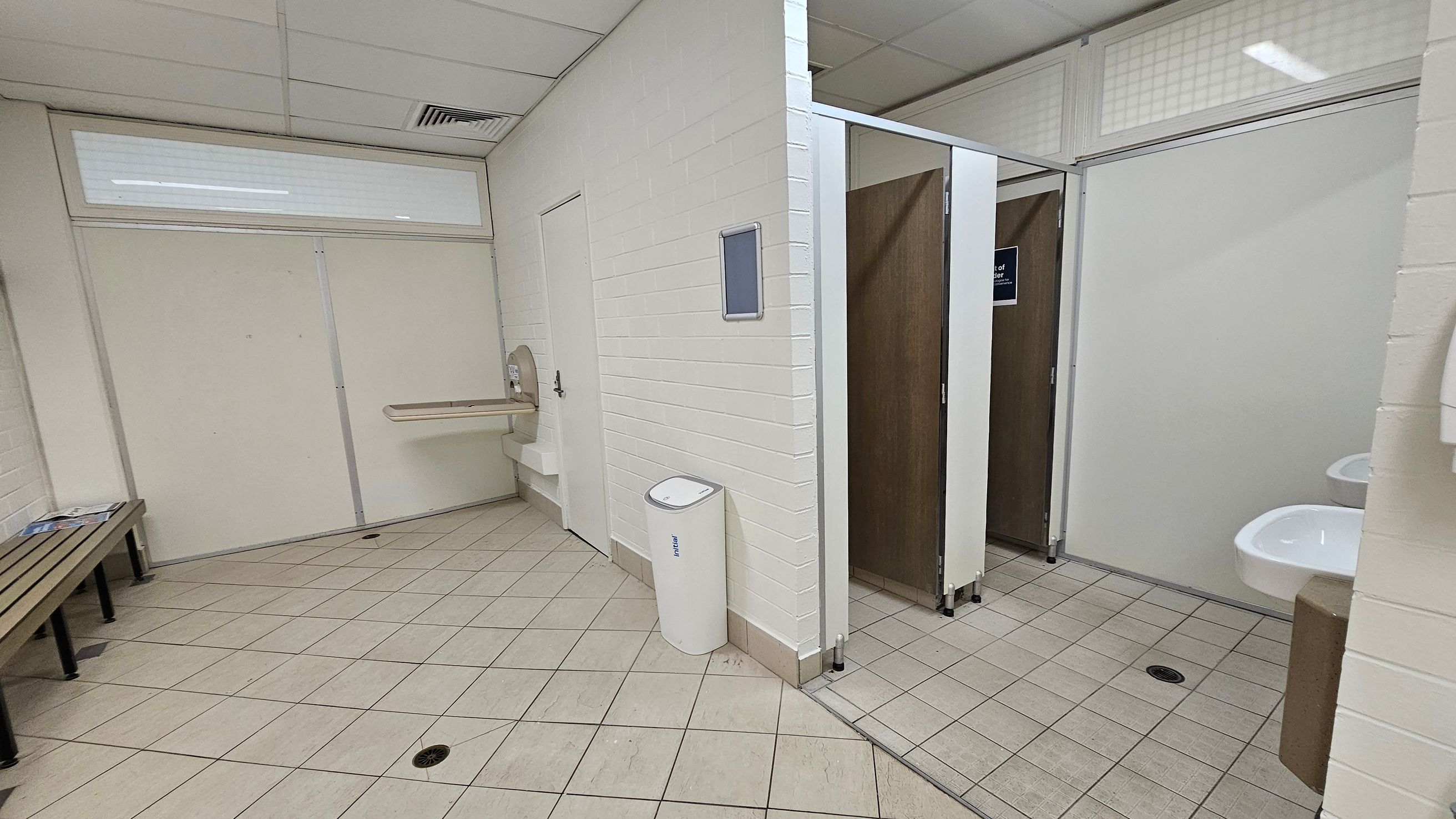 A public restroom with light green and beige tiled flooring. On the left, there's a wooden bench with black metal legs. A baby changing station is mounted on a light green wall. In the centre, a white bin is placed against a white brick wall. On the right, there are two toilet cubicles with wooden doors, one slightly open. A white sink is visible near the cubicles. The ceiling is white with fluorescent lighting and ventilation panels.