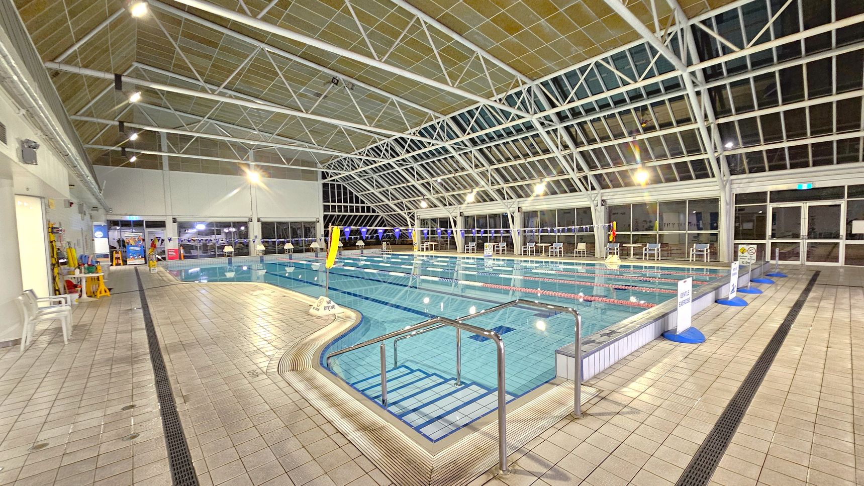 An indoor swimming pool with clear blue water, divided into lanes by floating ropes. On the left, there are white plastic chairs and cleaning equipment against a white wall. The right side features large glass windows and doors, with several white chairs and tables along the poolside. The ceiling is high with a metal framework and overhead lights. The floor is tiled in a light colour with drainage grates along the edges. Signs are placed around the pool area.
