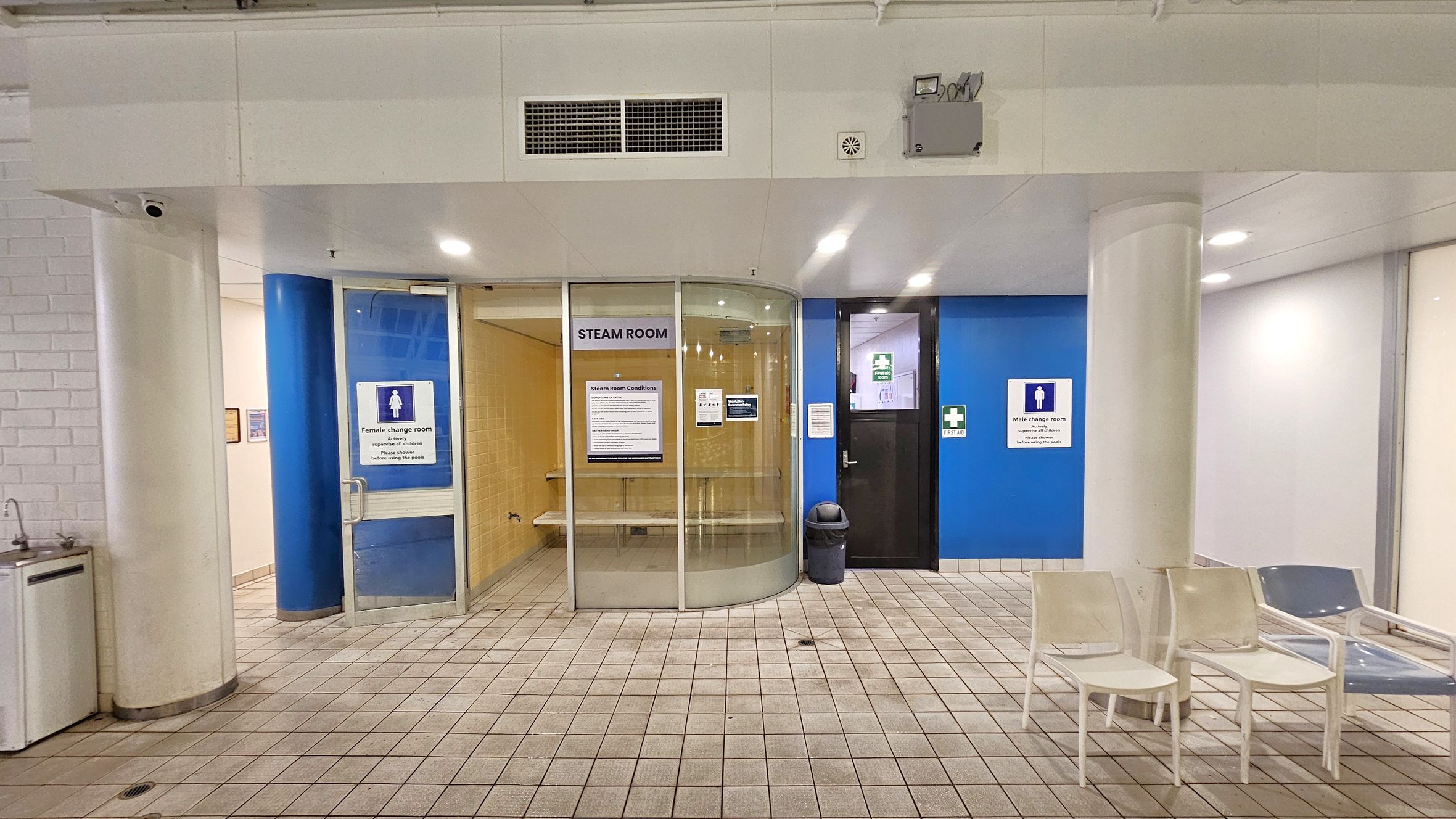 A tiled floor leads to a steam room with a glass door in the centre, displaying a sign with conditions. To the left, a door marked "Female change room" is set against a blue wall. On the right, a door labelled "Male change room" is also against a blue wall. White chairs are placed near a column on the right. The ceiling is white with lights and ventilation. A small sink is visible on the far left.