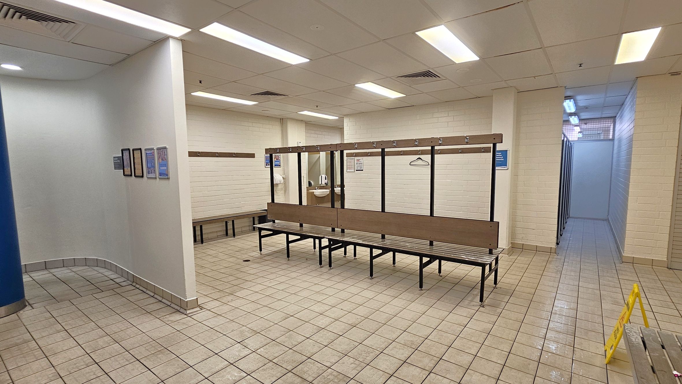 A changing room with tiled flooring and a grid-patterned ceiling with fluorescent lights. In the centre, there are benches with hooks above them. On the left, a curved wall with framed notices. On the right, a corridor leads to shower stalls. A yellow caution sign is on the floor. The walls are white brick, and a sink with a mirror is visible in the background.