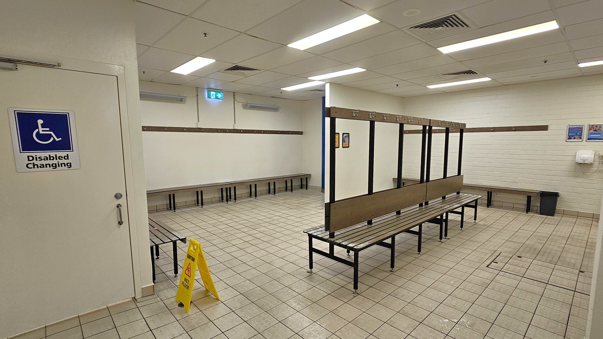 A changing room with tiled flooring and a suspended ceiling with fluorescent lights. On the left, a door marked "Disabled Changing" is visible. In the centre, there are benches with hooks above them for hanging clothes. The right side has more benches and a waste bin. A yellow caution sign indicating a wet floor is placed near the centre. The walls are light-coloured with a few posters and a hand dryer mounted on the right wall.