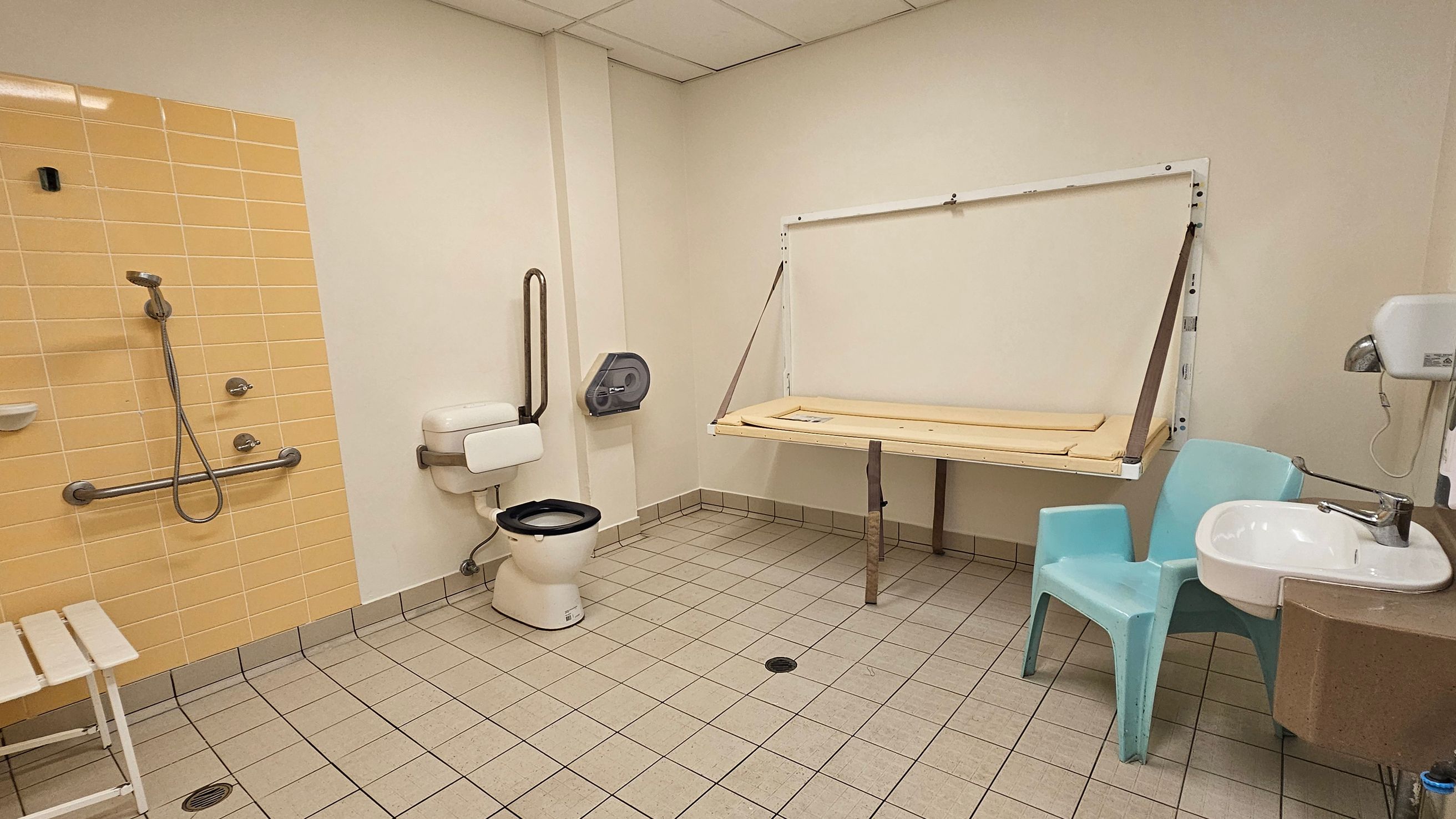 A bathroom with a tiled floor and ceiling panels. On the left, there is a shower area with yellow tiles, a handheld showerhead, and a grab bar. A white bench is nearby. In the centre, a toilet with a grab bar and a wall-mounted paper dispenser is visible. On the right, a foldable changing table is mounted on the wall. A light blue plastic chair is next to a white sink with a tap, and a hand dryer is mounted above the sink.