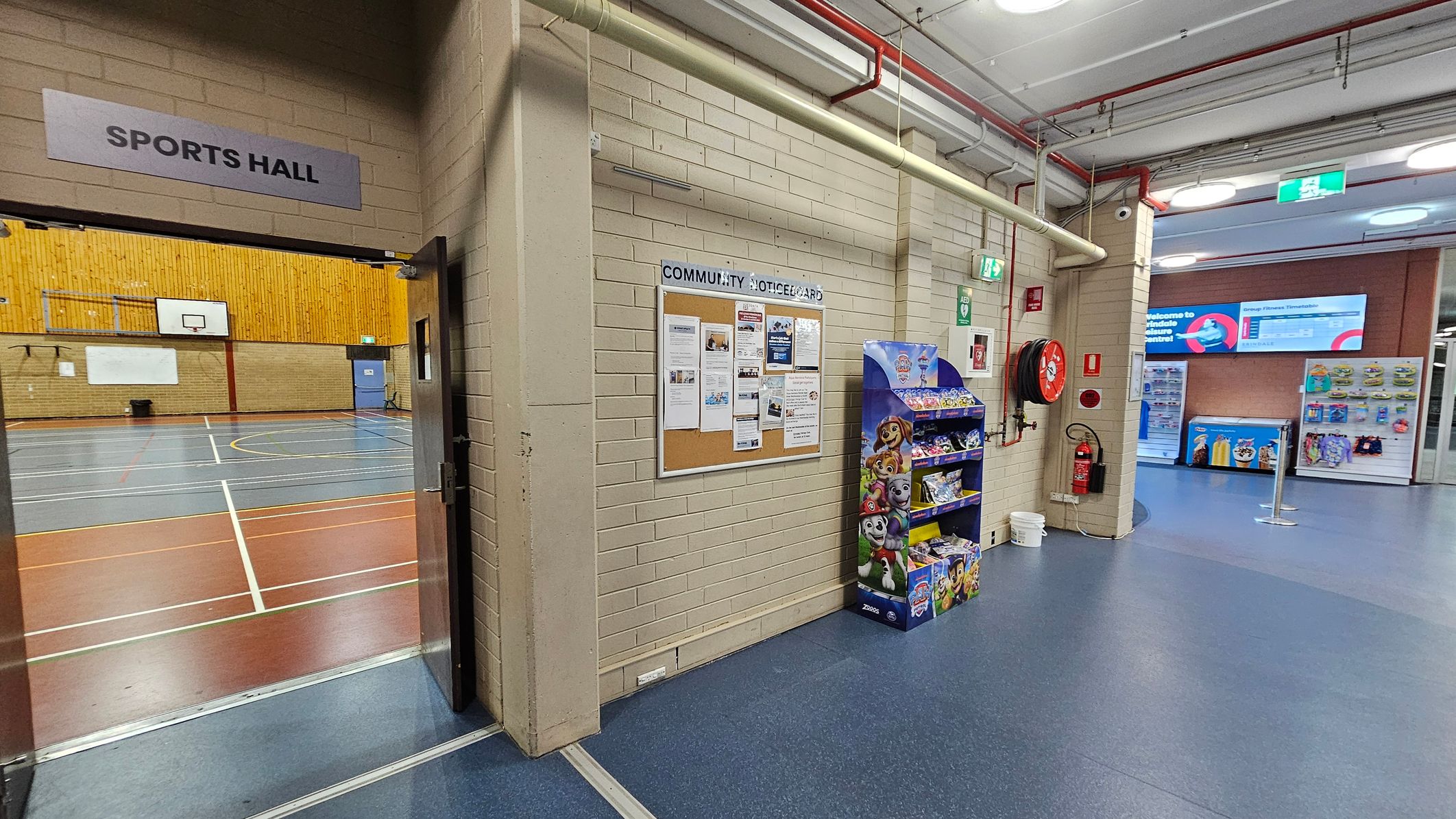 On the left, an open door leads to a sports hall with a wooden wall and basketball hoops. In the centre, a community noticeboard is mounted on a brick wall, with various papers pinned to it. Below, a display stand with children's snacks is visible. On the right, a fire hose reel and extinguisher are mounted on the wall. The floor is blue, and the ceiling has exposed pipes and lights. In the background, vending machines and a digital sign are visible.